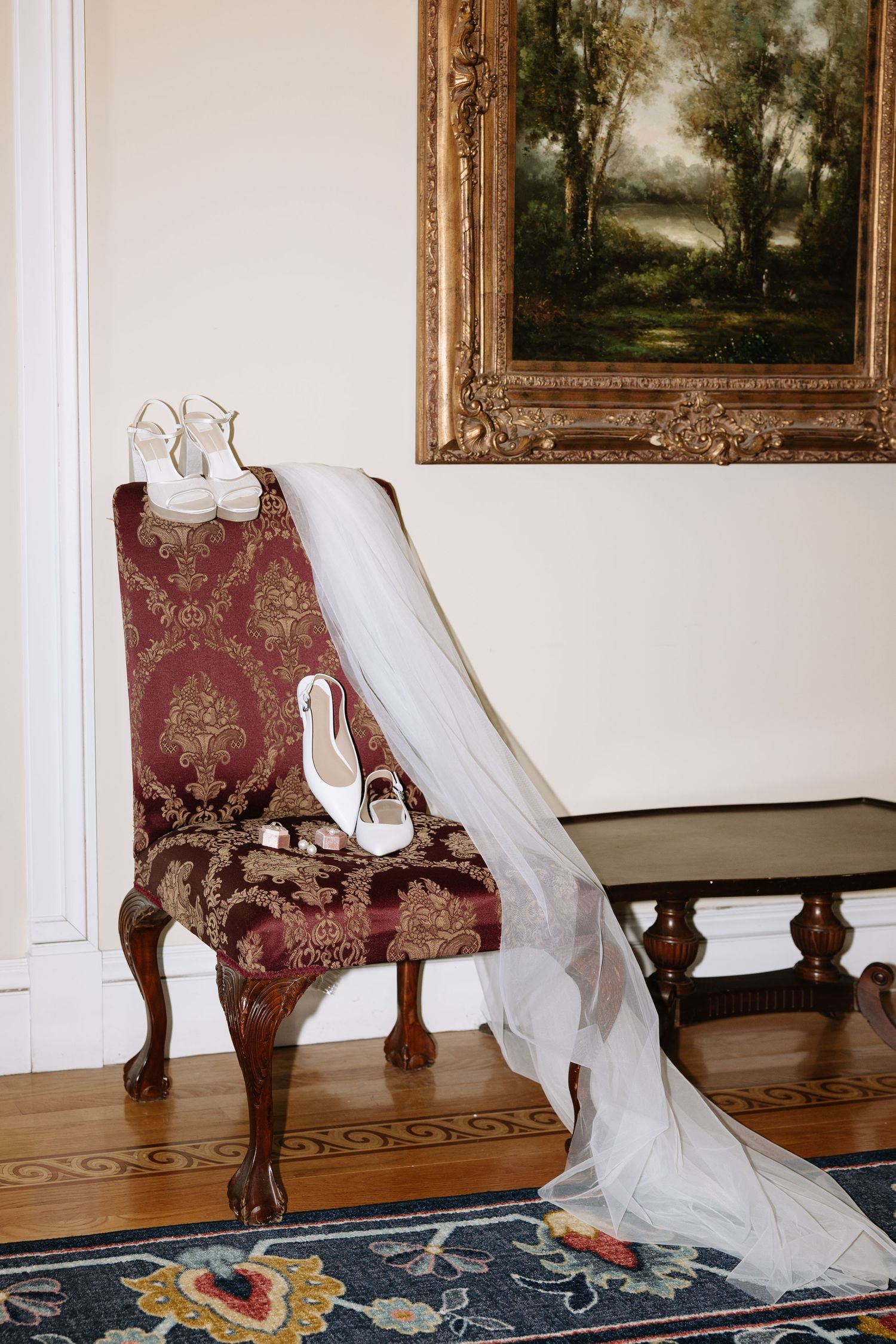 Wedding shoes and veil artistically arranged on antique furniture beside classical paintings.