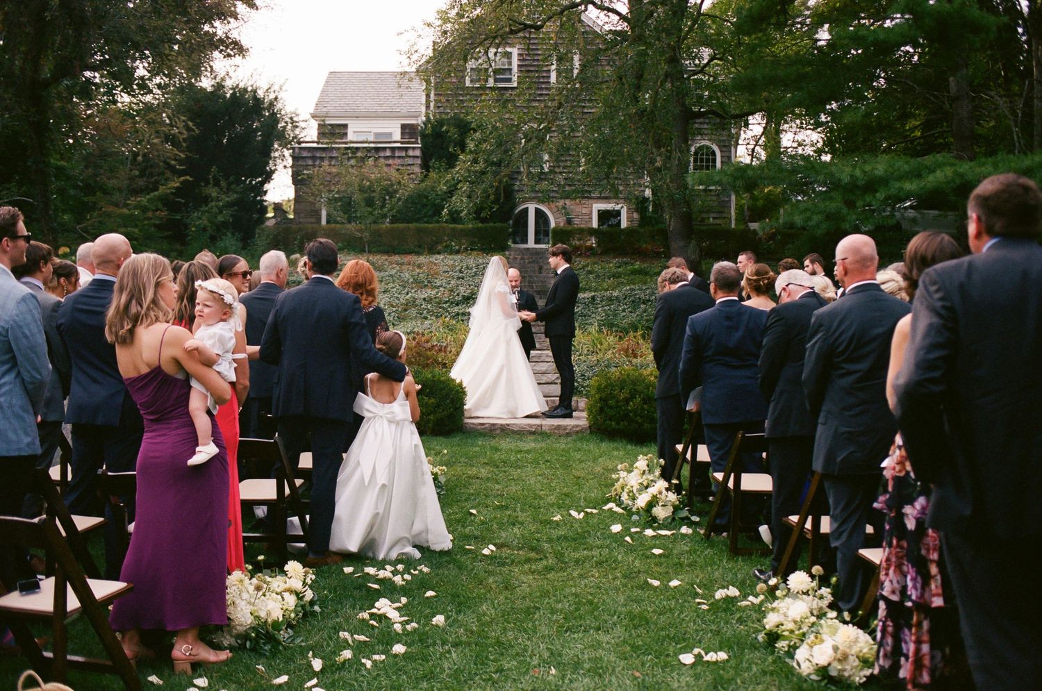Outdoor wedding ceremony setup with guests seated on wooden chairs and white flower petals lining the aisle.