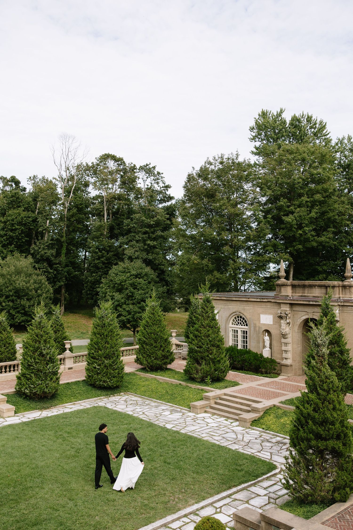 A couple walking near a classical stone building surrounded by manicured gardens.