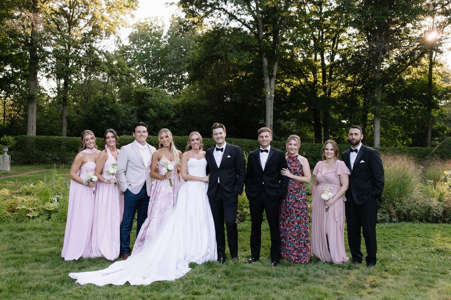 Wedding party in pink dresses and black tuxedos poses together on garden grounds.