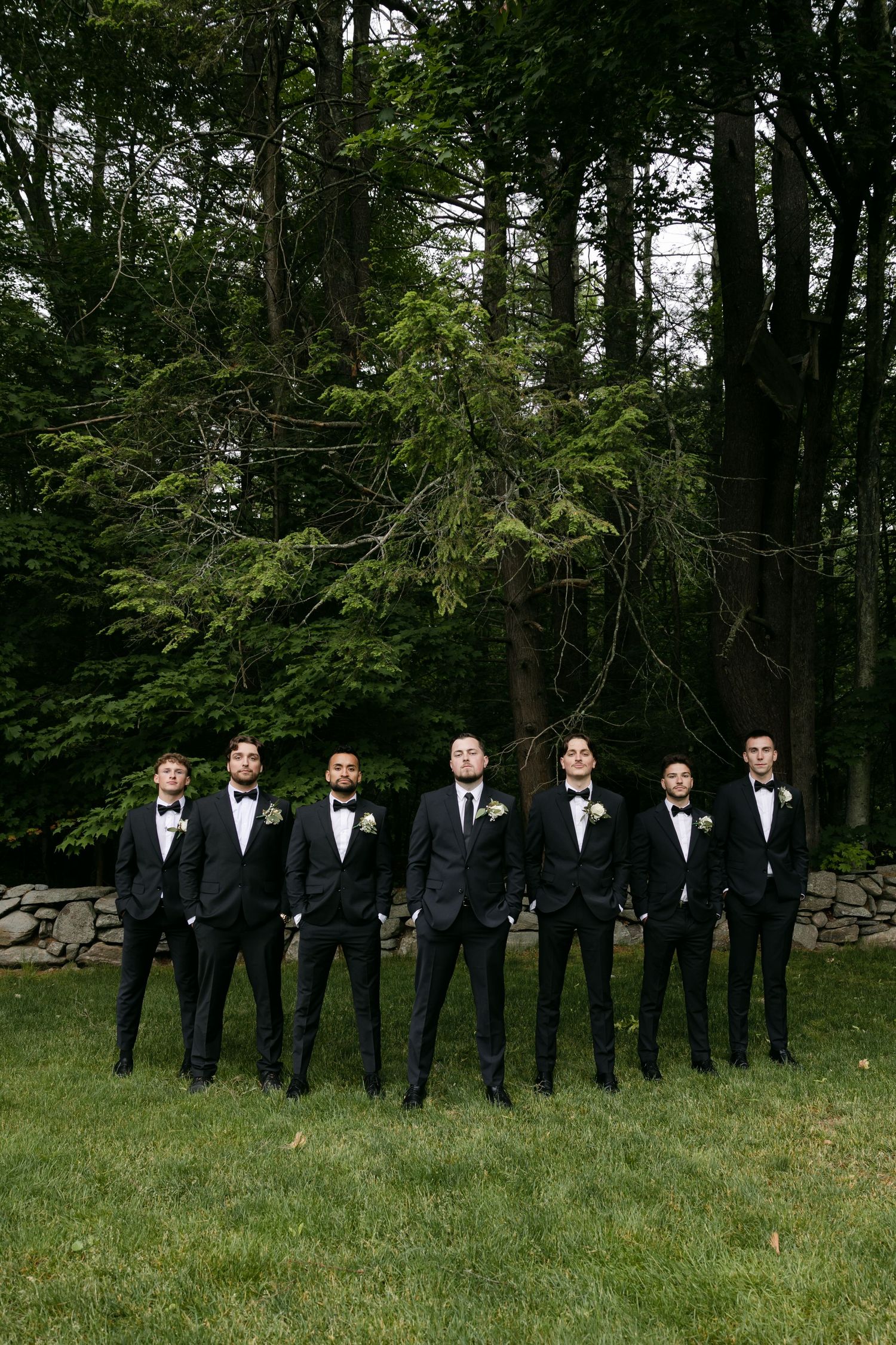 Groomsmen in black tuxedos line up on grass in front of wooded area.