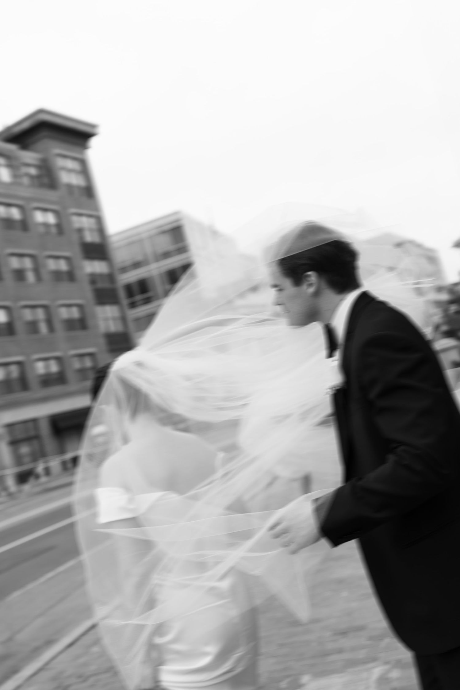 Black and white photo of a Jewish bride and groom walking outdoors, her veil blowing in the wind.