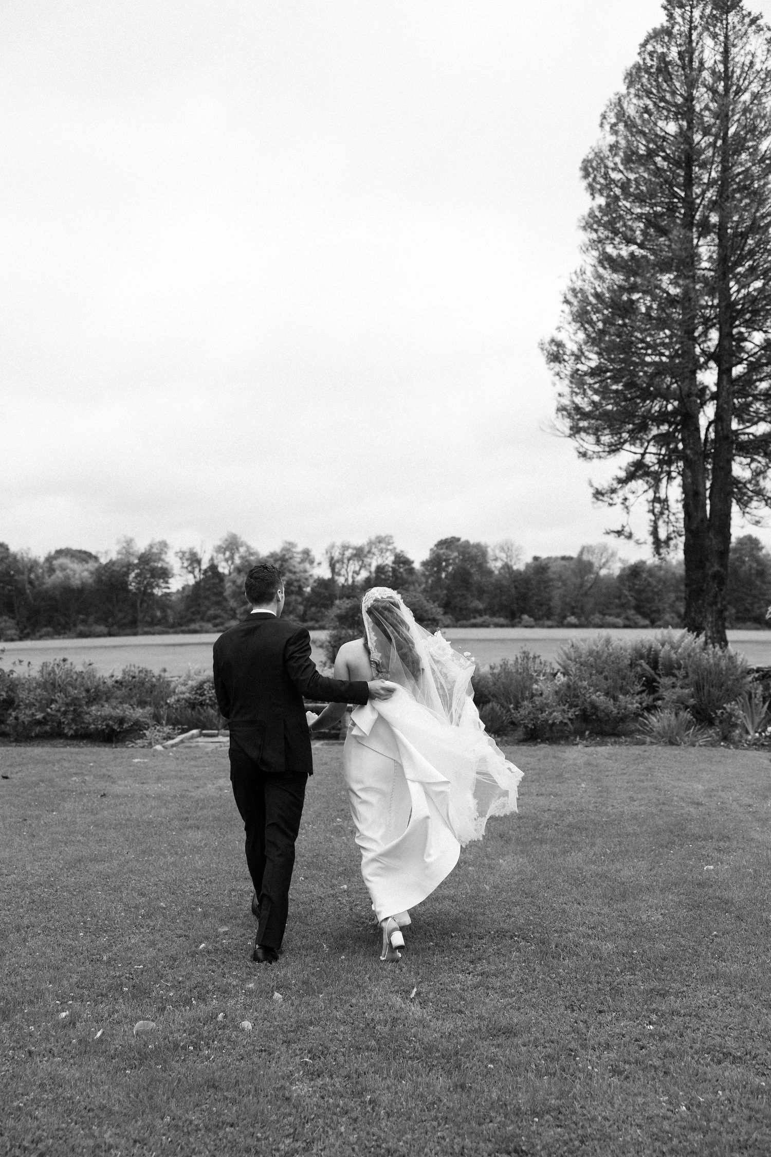 Black and white photo of bride and groom walking hand in hand across lush green lawn on their wedding day.