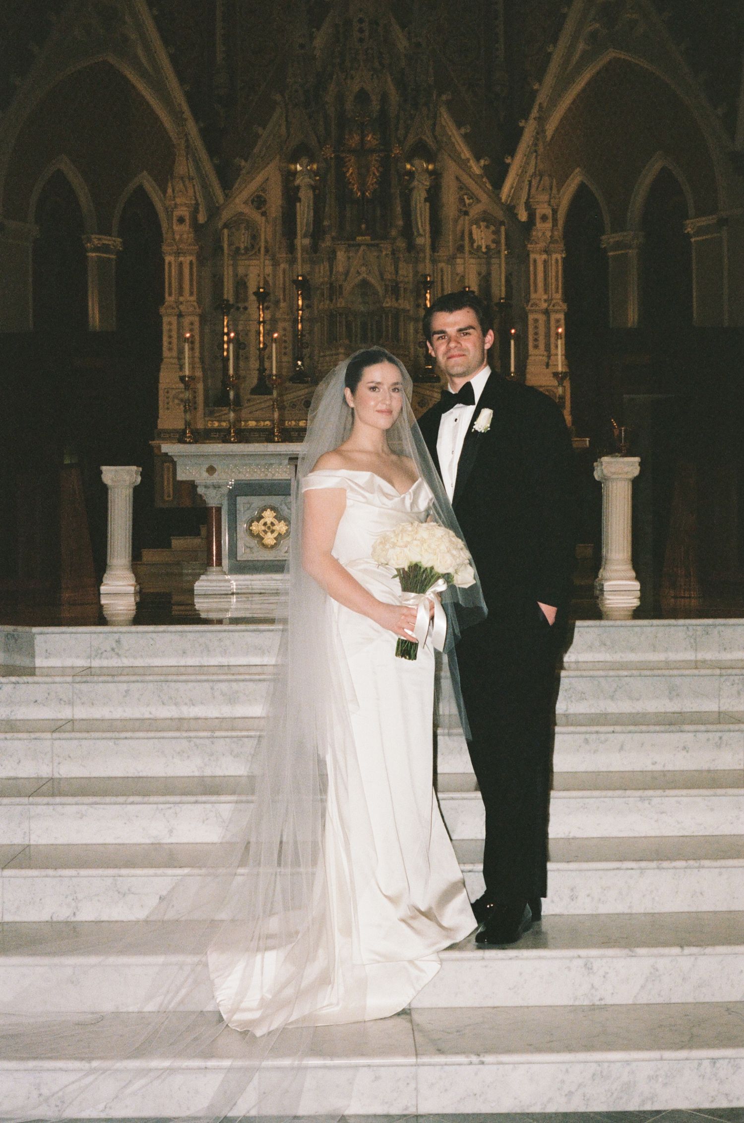 Bride and groom posing on church altar steps, bride in white gown with veil holding bouquet, groom in tuxedo.