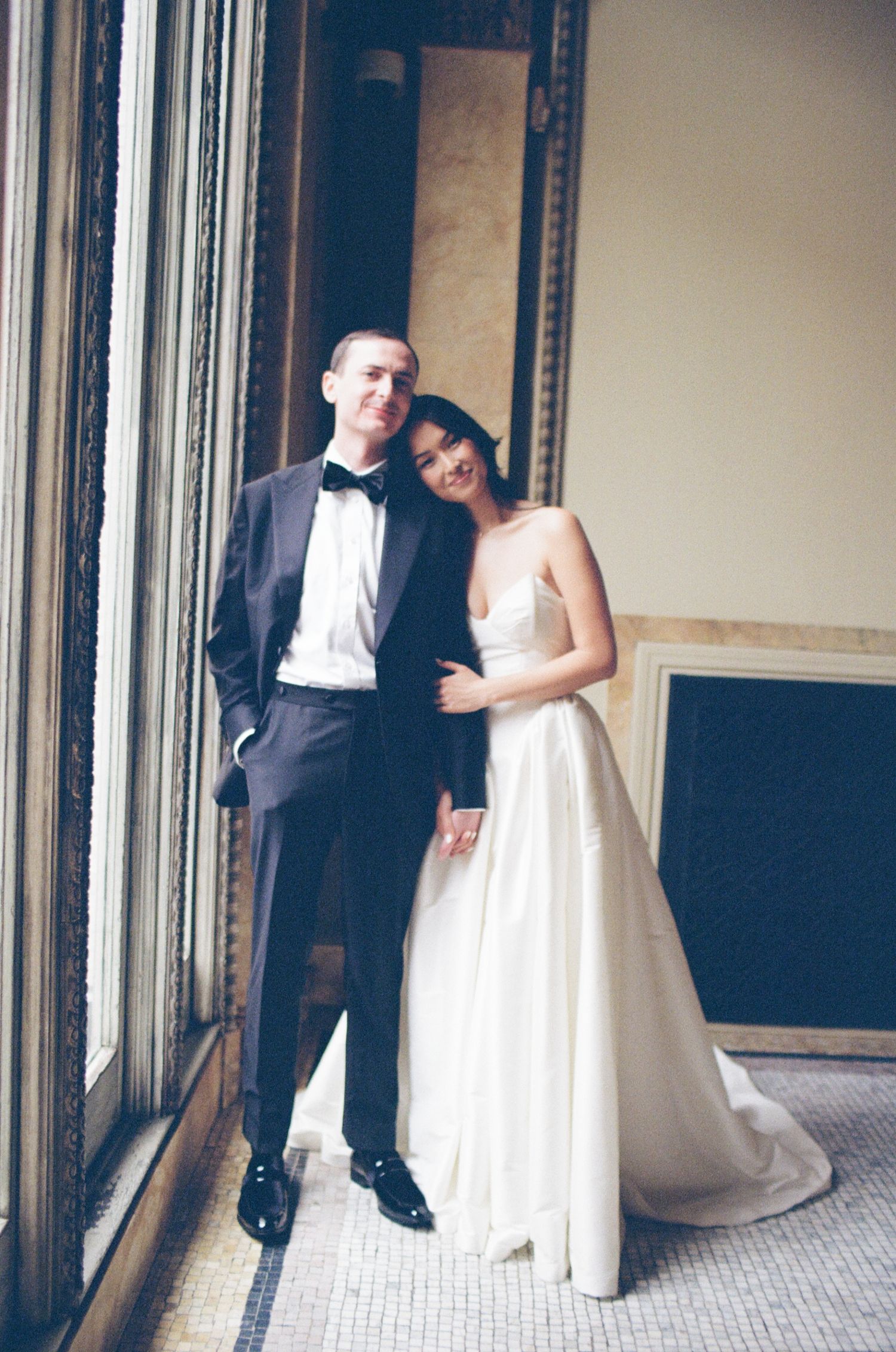 Elegant bride in white ball gown and groom in black tuxedo posing together near large windows at wedding venue.