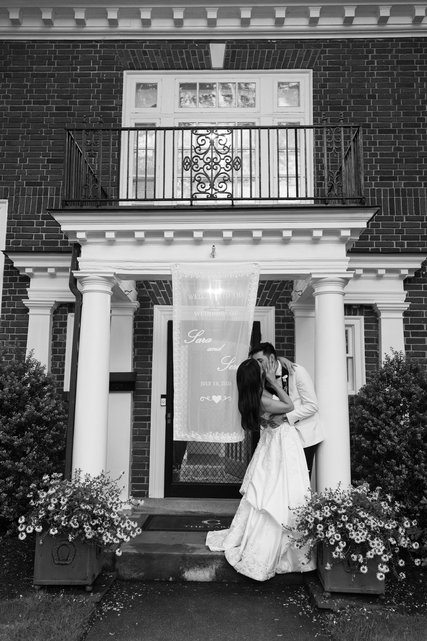 Bride and groom share a romantic kiss on the steps of a classic brick colonial mansion adorned with white columns.