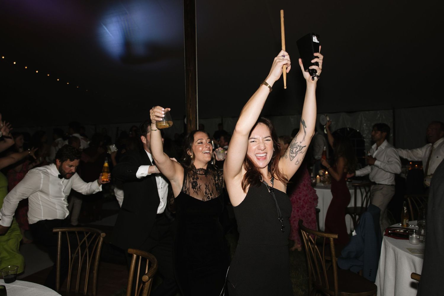 Two women in black dresses dancing and celebrating at an indoor wedding reception party at night.