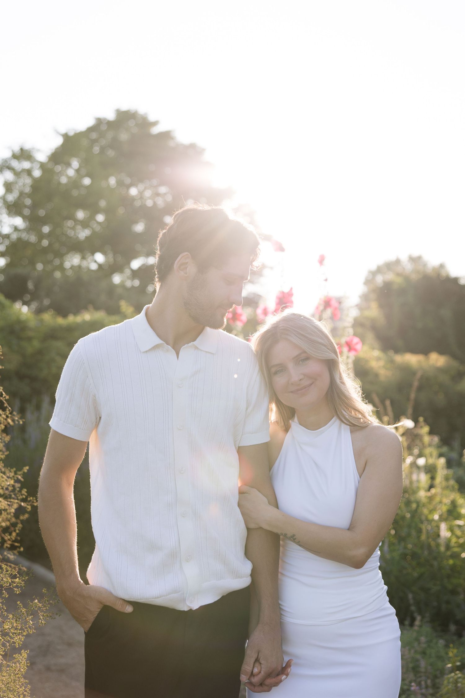 Couple in white outfits embracing in a sunlit garden with pink flowers and green trees in the background.