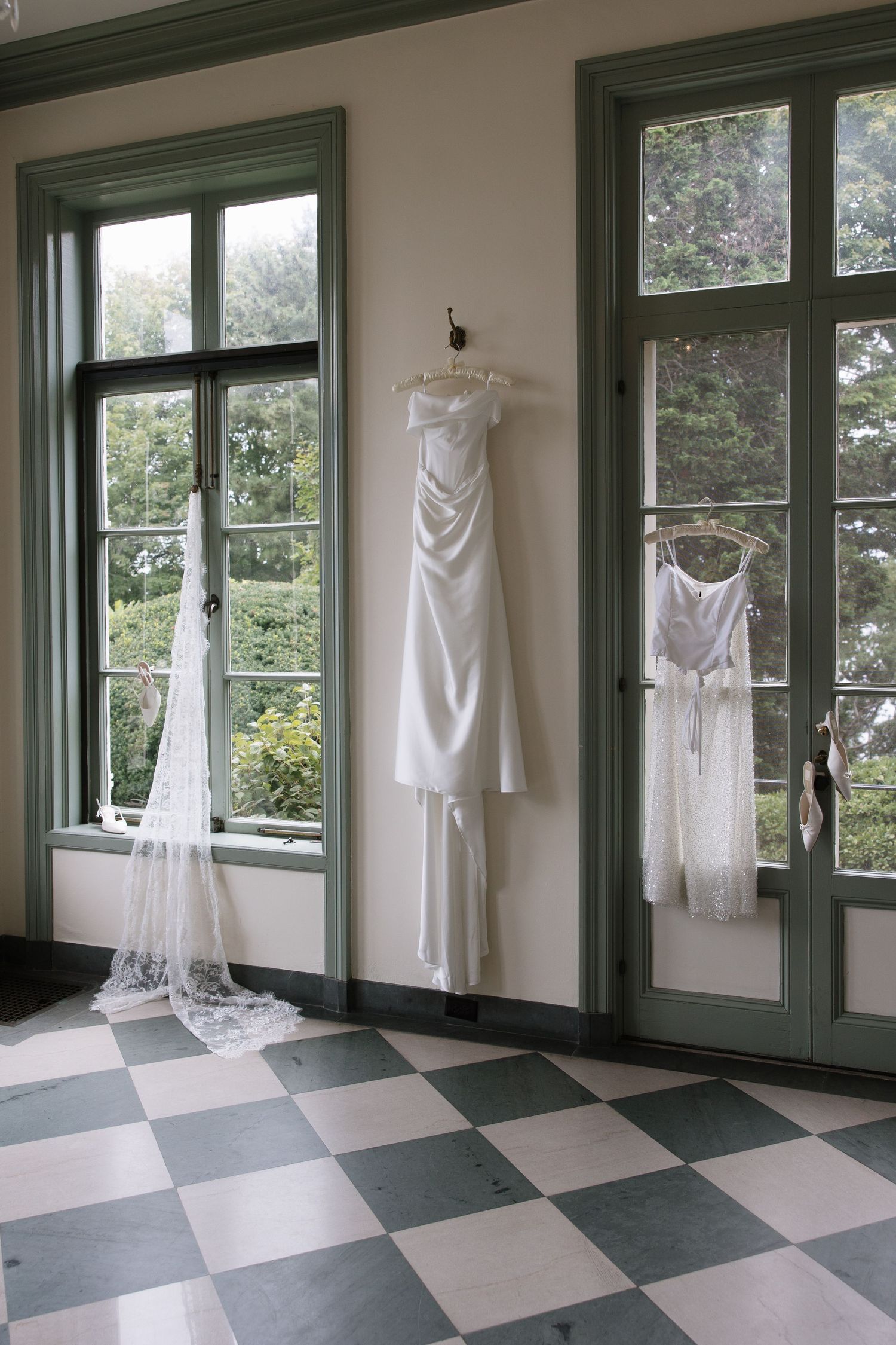 White wedding dress and veil hanging on wall between green-framed windows with checkered floor in elegant room.