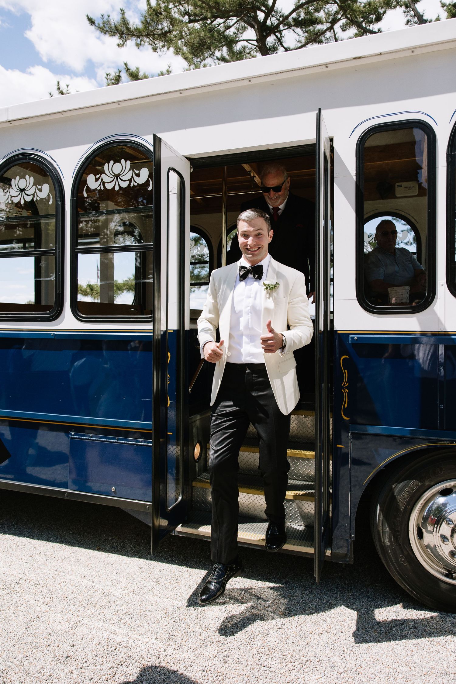 Groom in white tuxedo jacket and bow tie steps off a blue and white trolley bus giving a thumbs up.