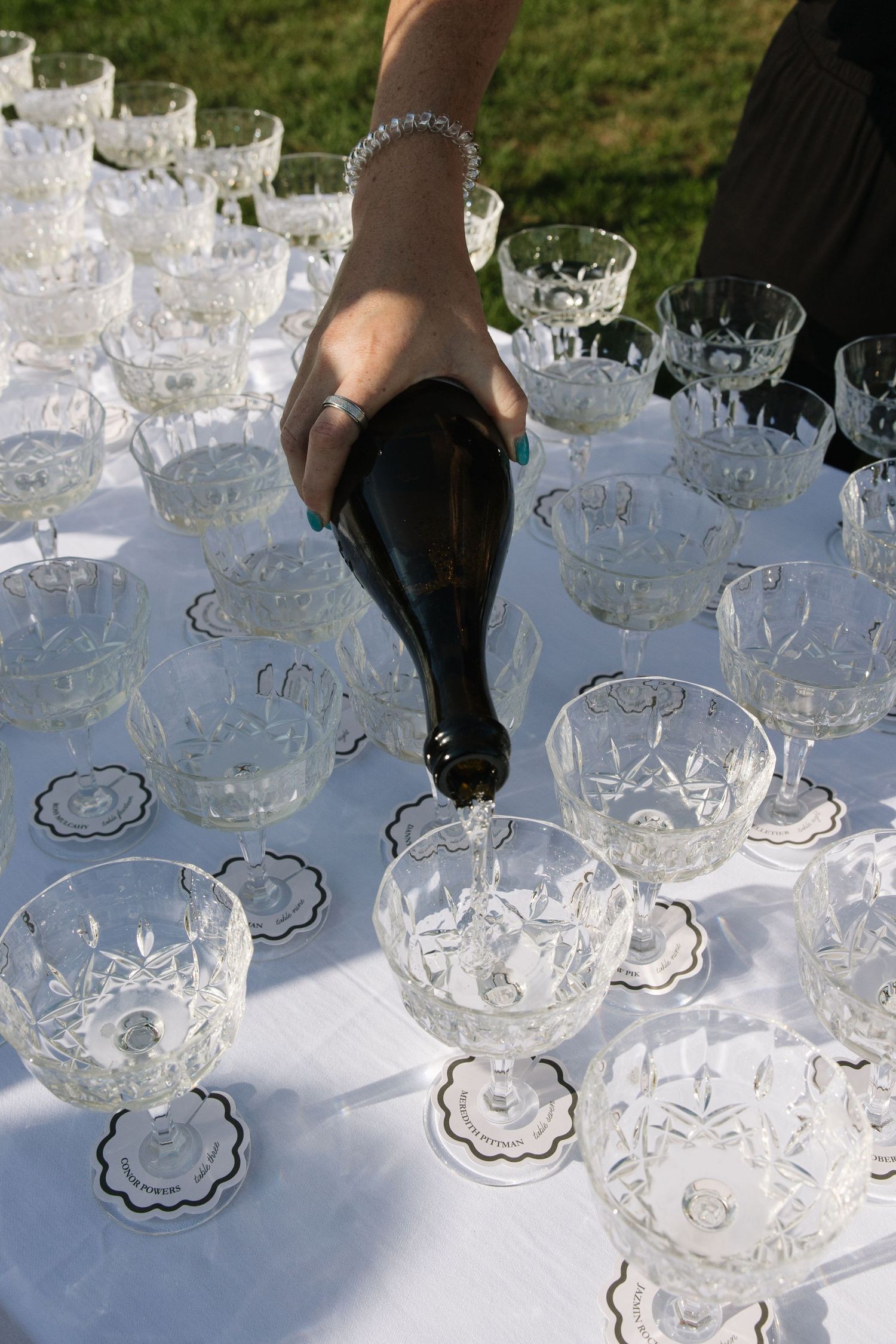 Hand pouring champagne from dark bottle into crystal glasses on white tablecloth at outdoor event.