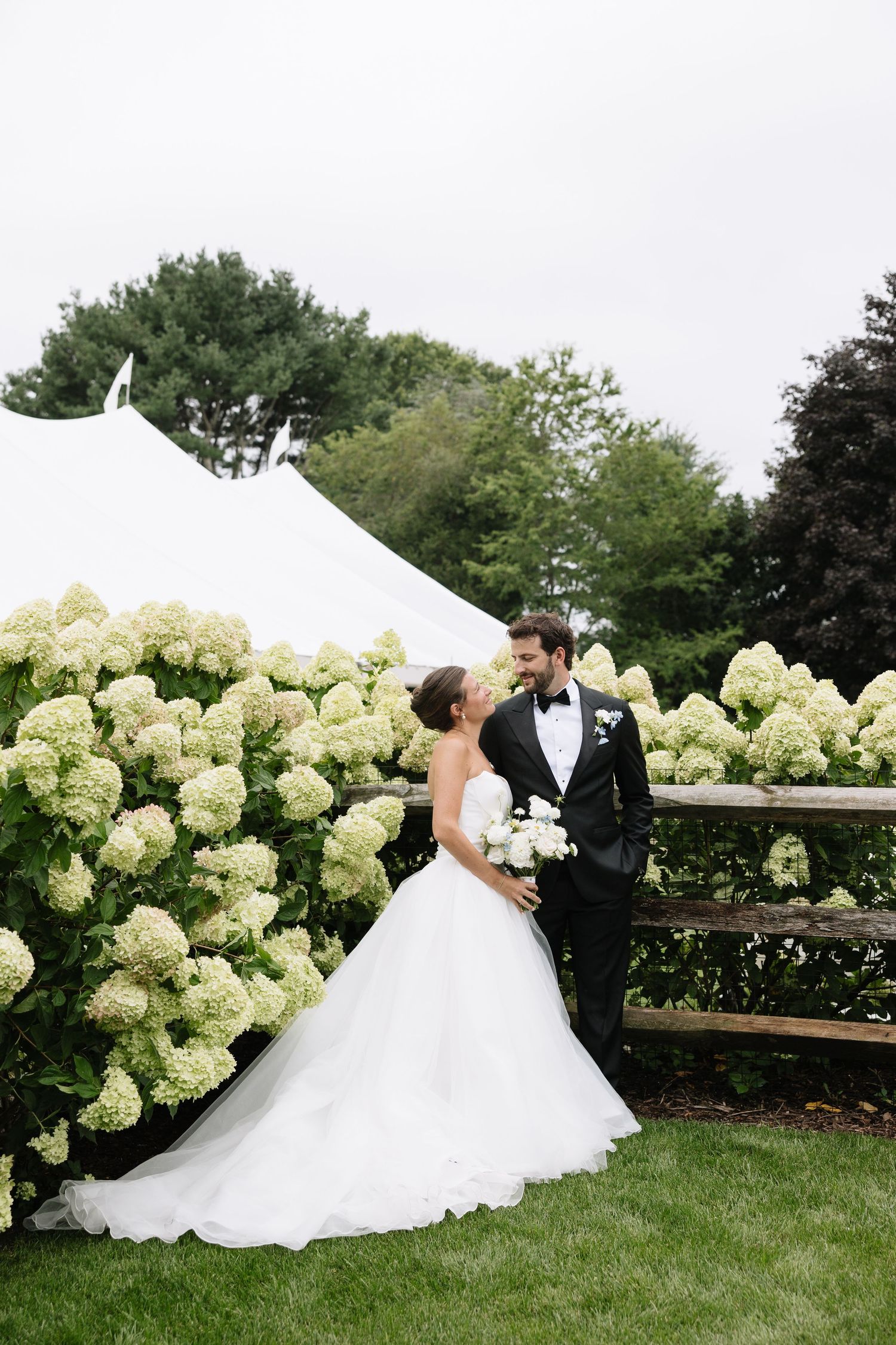 Bride and groom share a romantic moment beside white hydrangeas near a wedding tent outdoors.