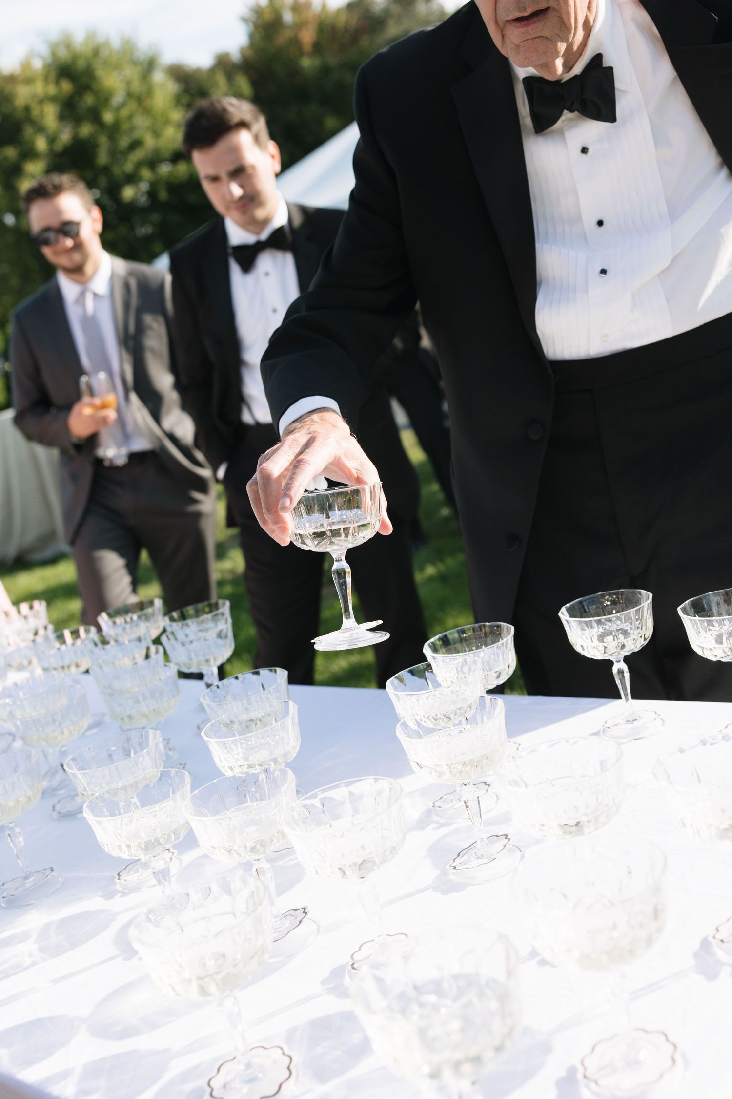 Elegantly dressed man in tuxedo picks up crystal champagne coupe from white tablecloth at outdoor wedding reception.