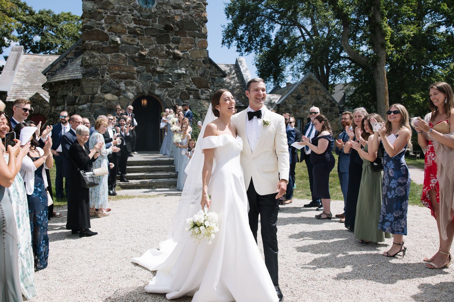 Bride and groom walk through cheering guests outside stone church, bride in off-shoulder gown holding white bouquet.