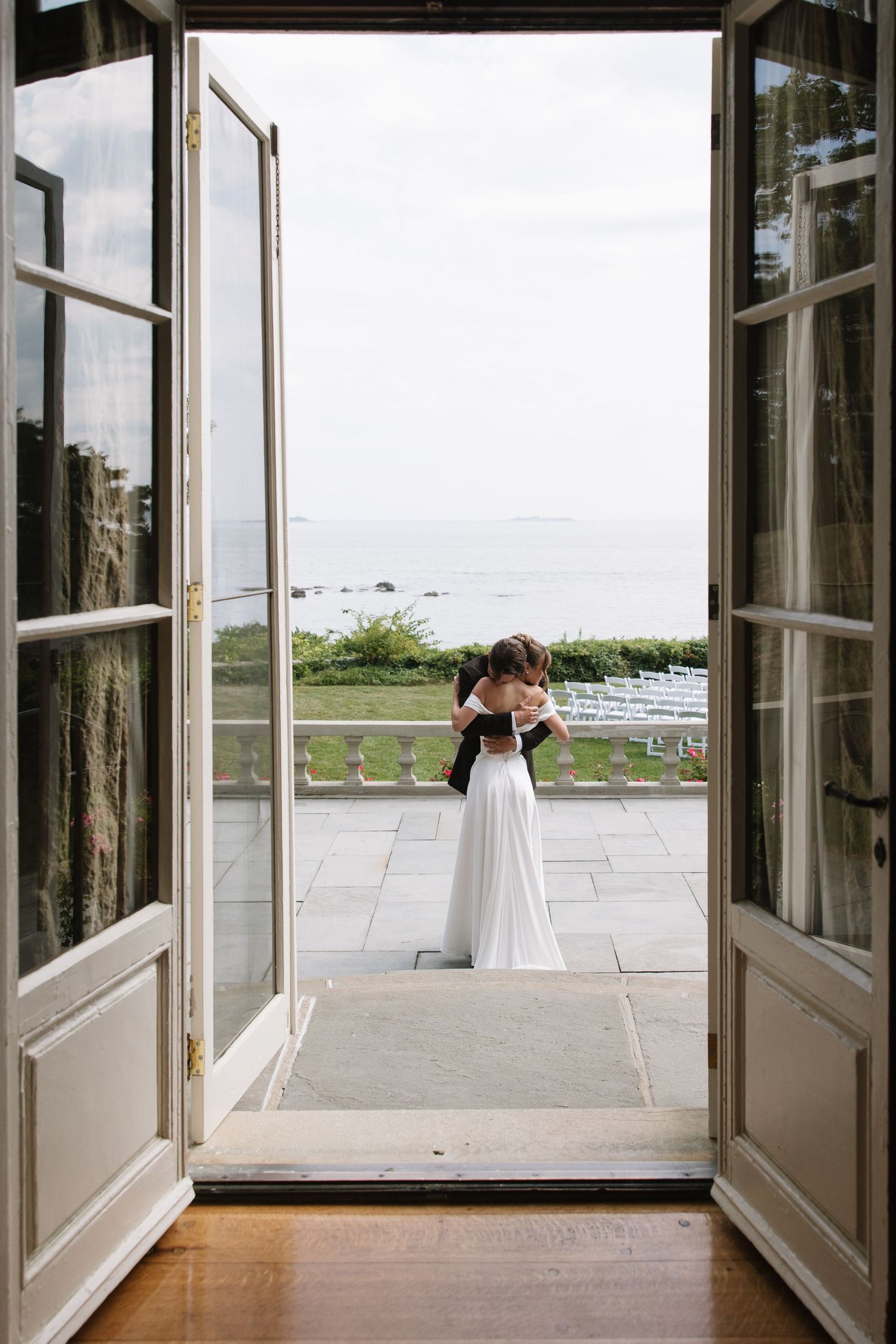 Bride and groom embrace on an elegant seaside terrace, viewed through open French doors with ocean views.