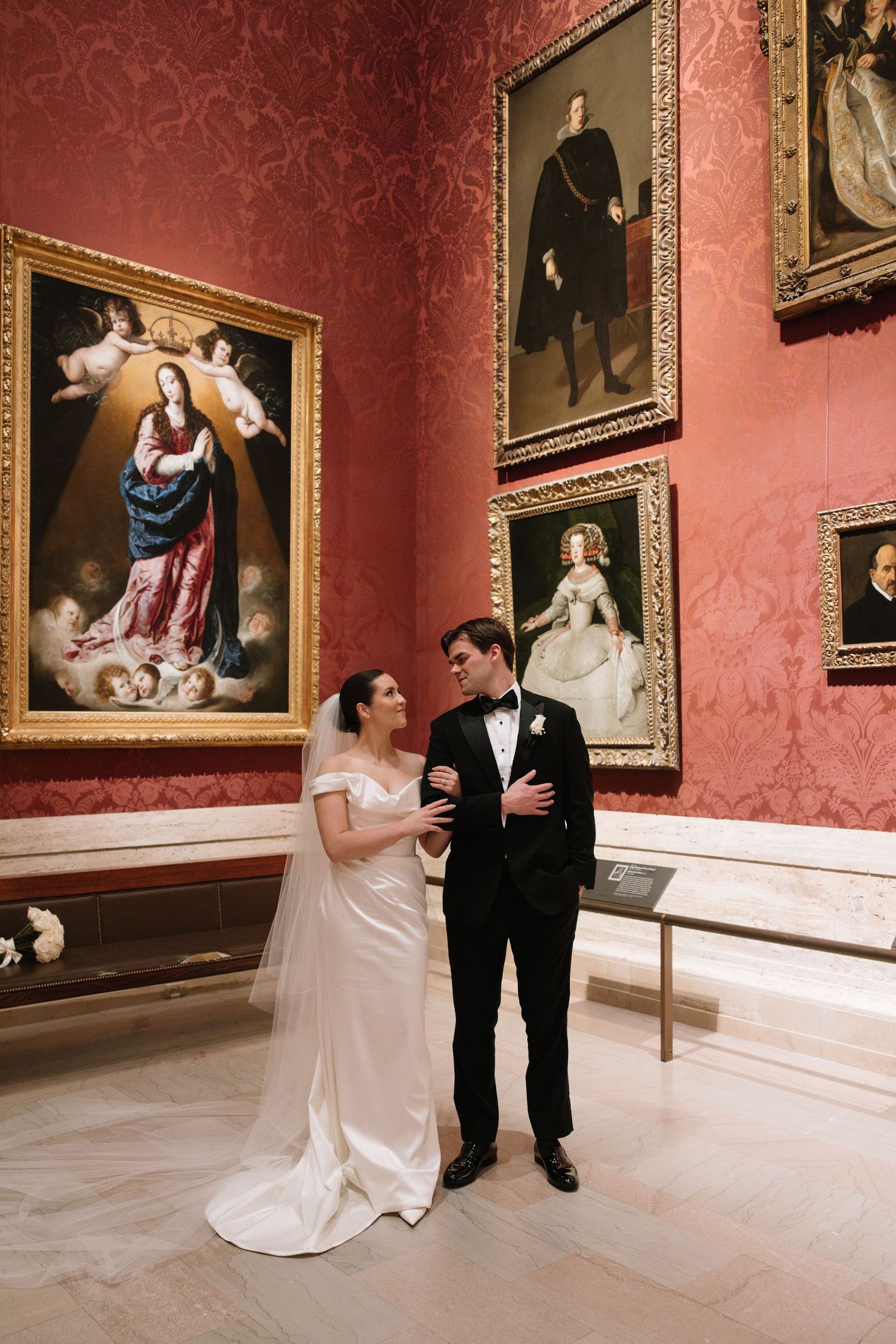 Elegant wedding couple sharing a romantic moment surrounded by old master paintings in a red museum gallery.