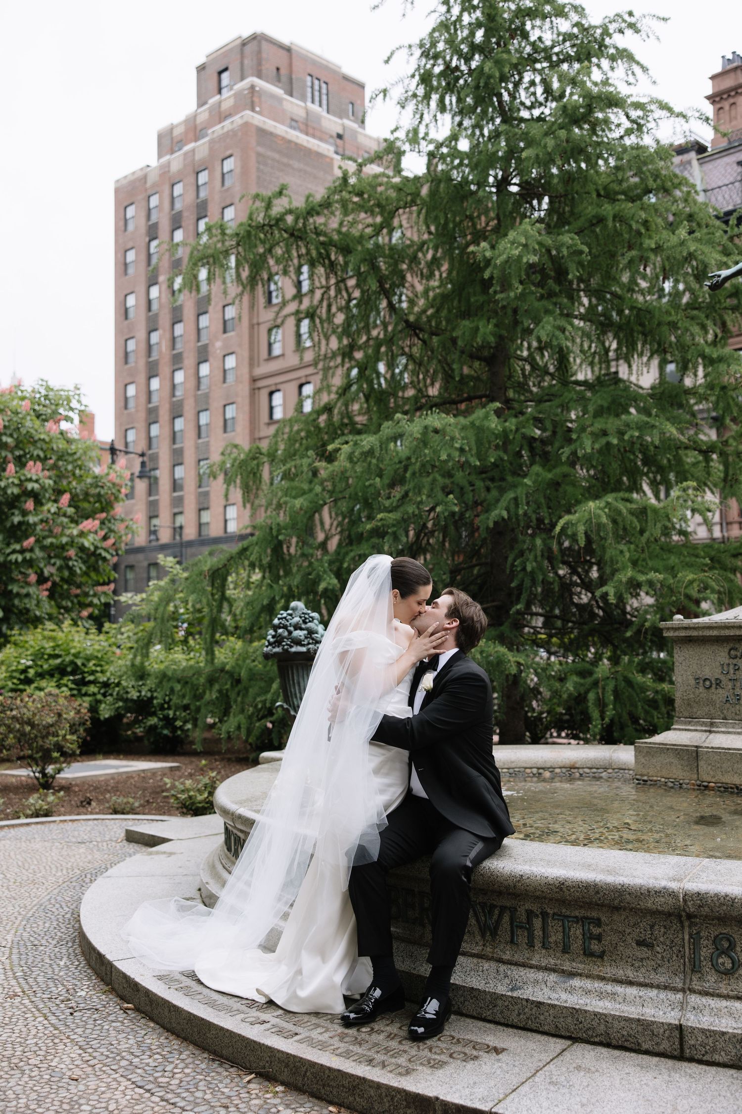 Bride and groom share romantic kiss seated at stone fountain in urban garden.
