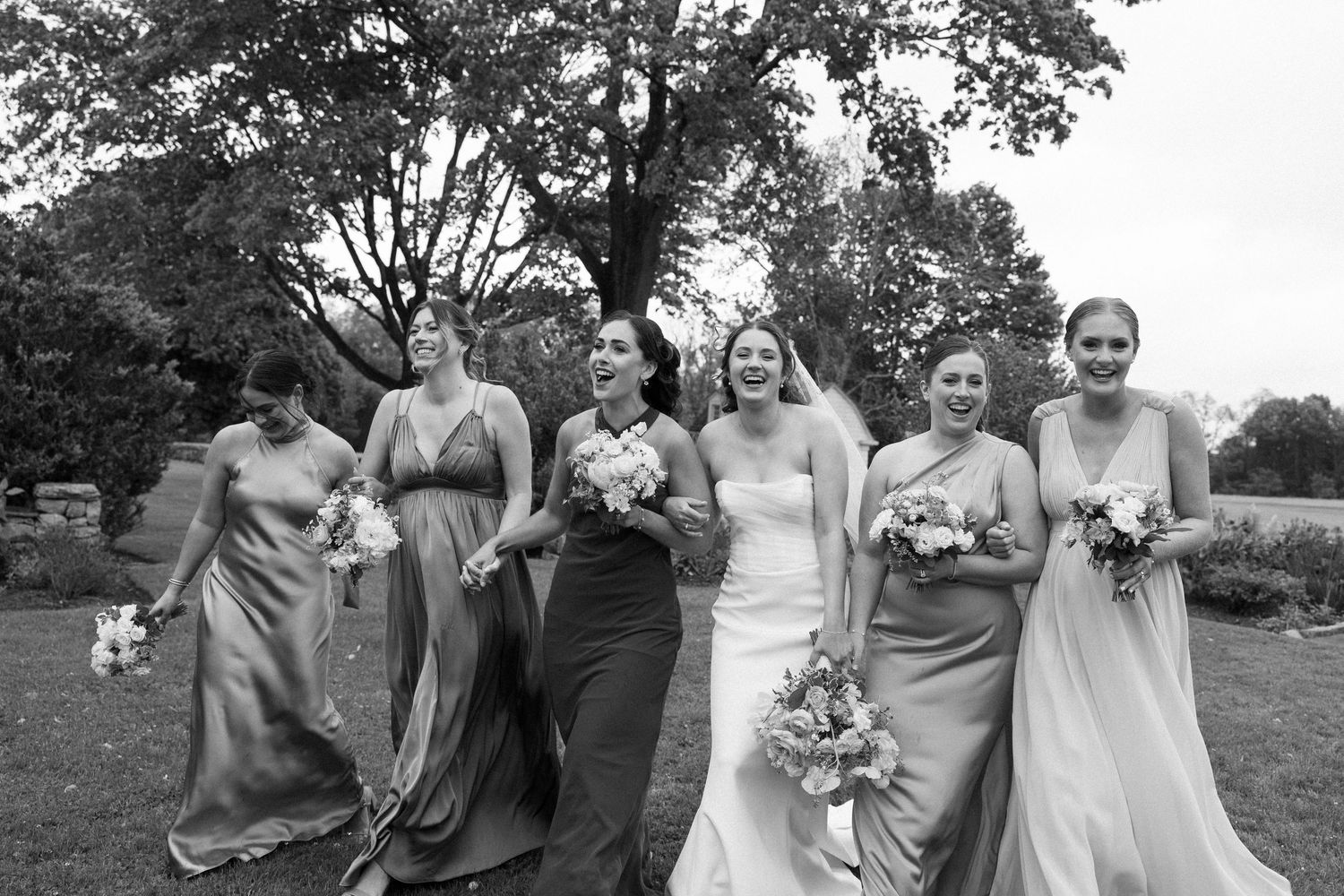 Black and white photo of a bride and five bridesmaids laughing and walking together outdoors at a wedding.