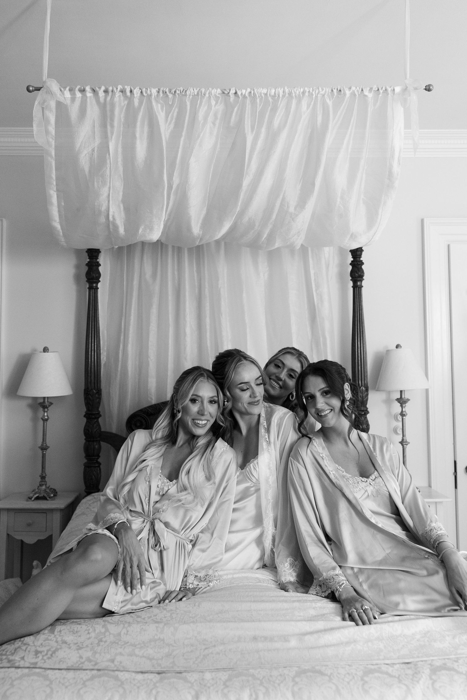 Four women in silk robes sitting together on a canopy bed, smiling and posing in a black and white bridal getting ready photo.