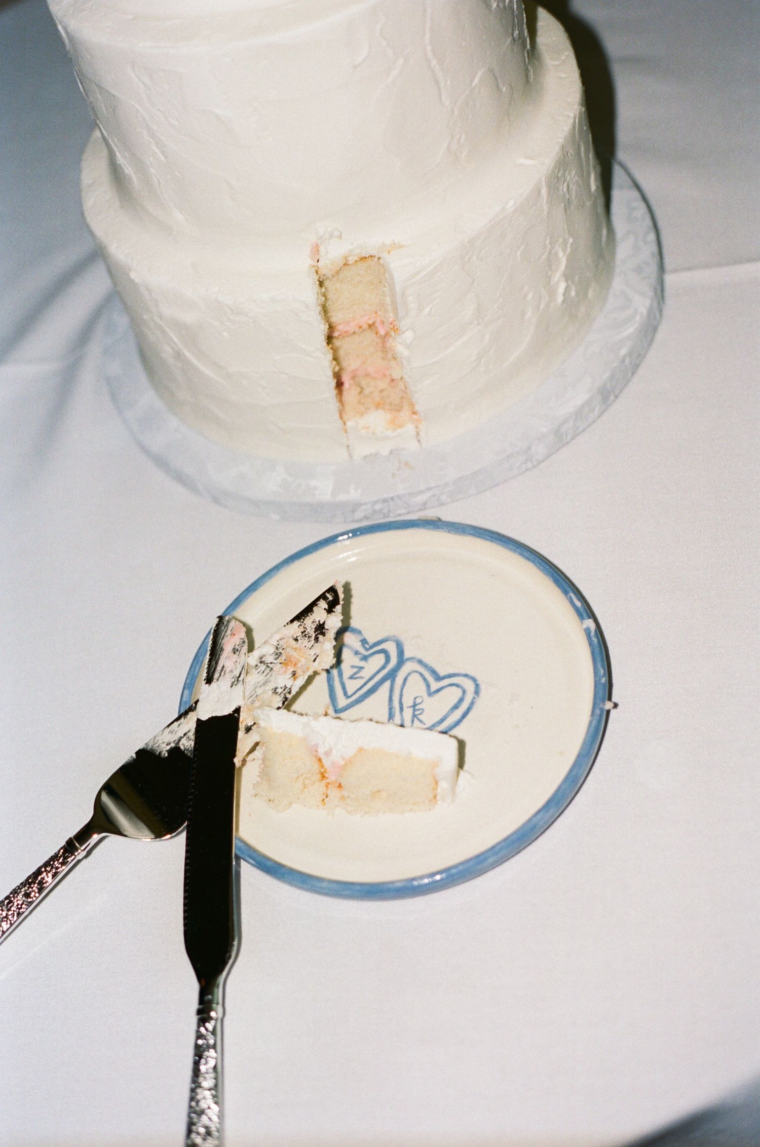 White two-tier wedding cake with a slice removed, served on a blue-rimmed plate with heart cake topper and silver serving knife.