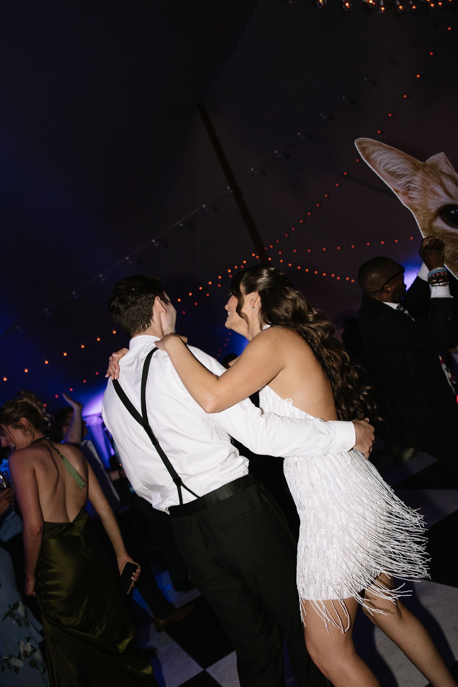 Couple dancing at prom in white fringe dress and suspenders under blue lights on checkered dance floor.