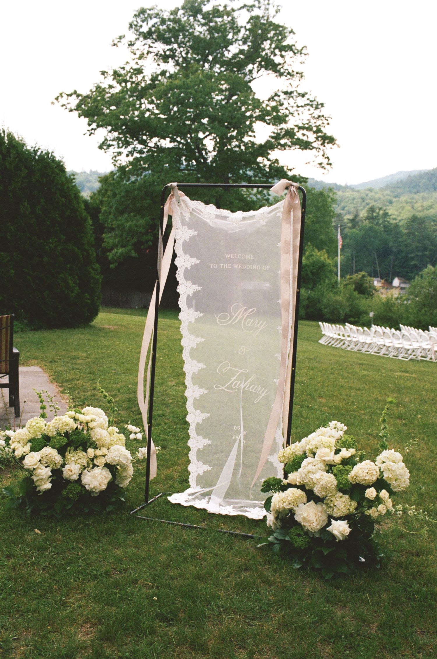 Outdoor wedding ceremony entrance with lace veil sign, white hydrangeas, and chairs on green lawn.