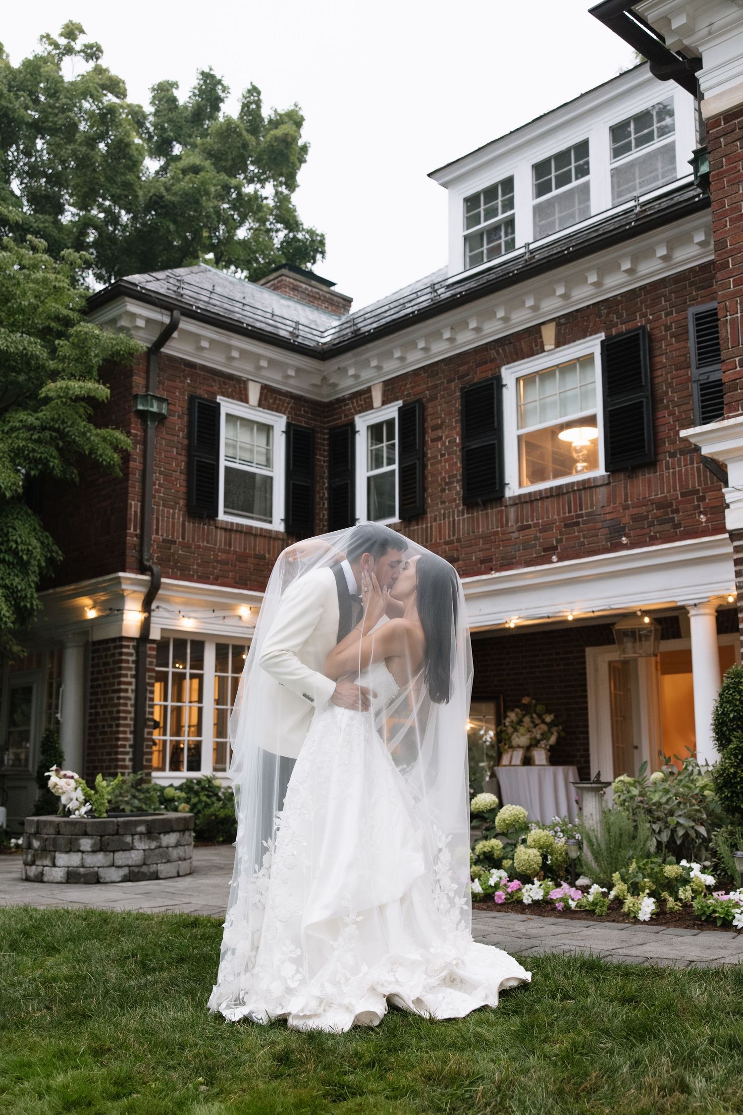 Bride and groom share a kiss under flowing veil in front of elegant brick manor house at dusk.