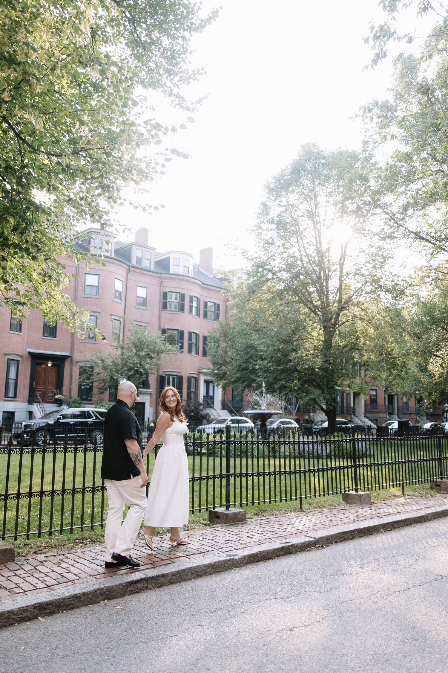 Couple walking along iron-fenced sidewalk in front of historic brick buildings.
