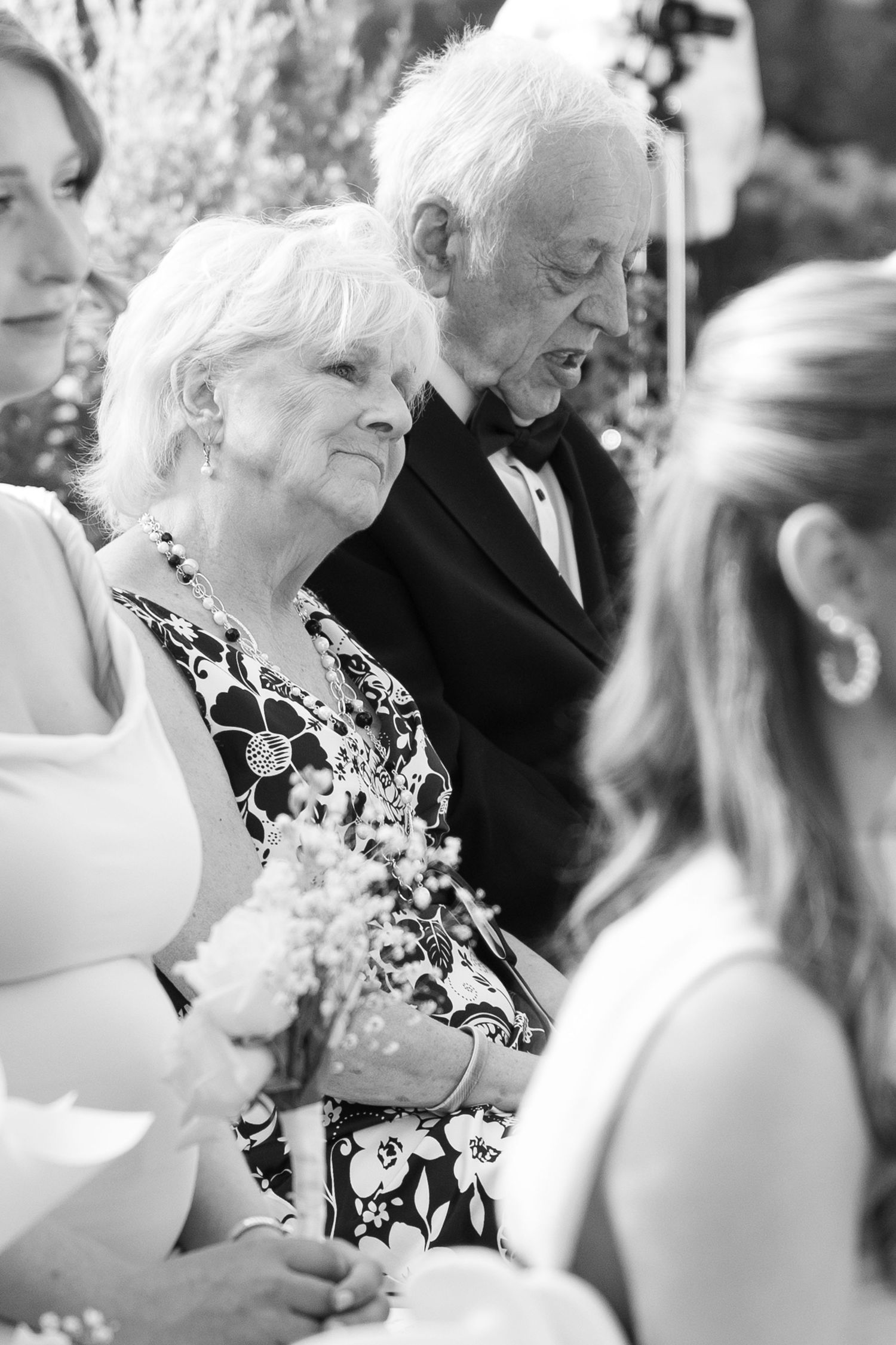 Black and white candid moment during a wedding ceremony with attendees holding flowers.