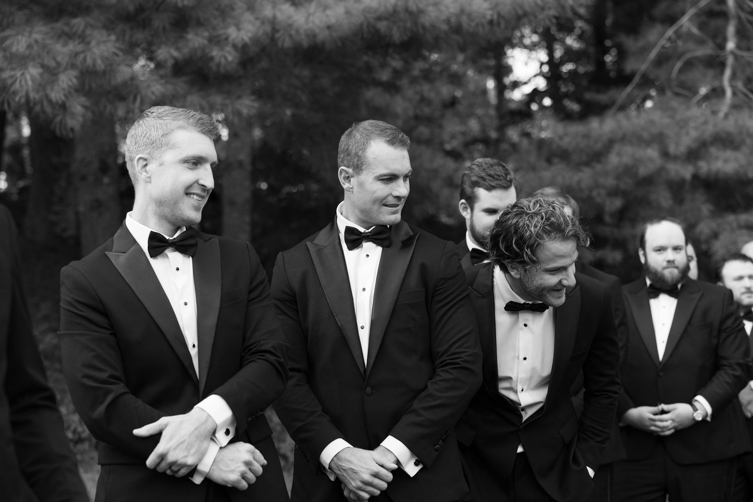 Groomsmen in black tuxedos share a candid moment during outdoor wedding ceremony.