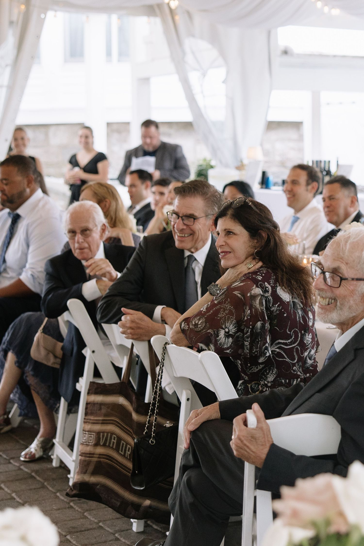 Wedding guests seated in white chairs under a draped tent during an outdoor ceremony.