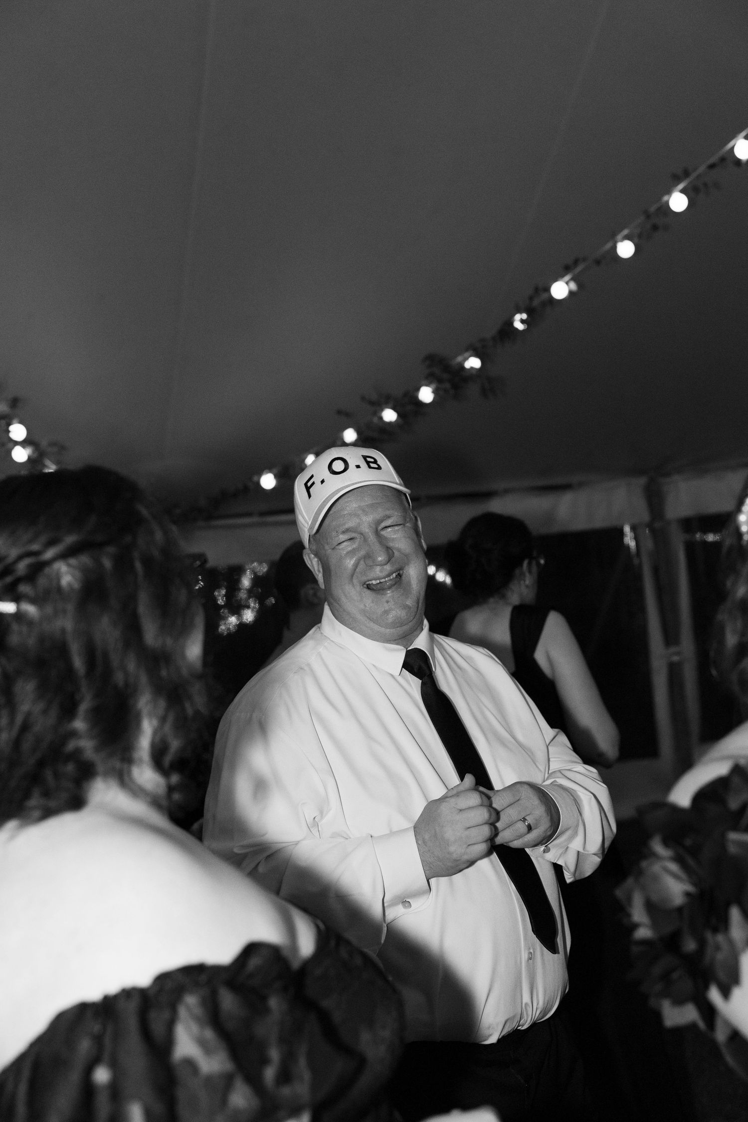 Man wearing F.O.B. hat laughing at a wedding reception under a tent with string lights.