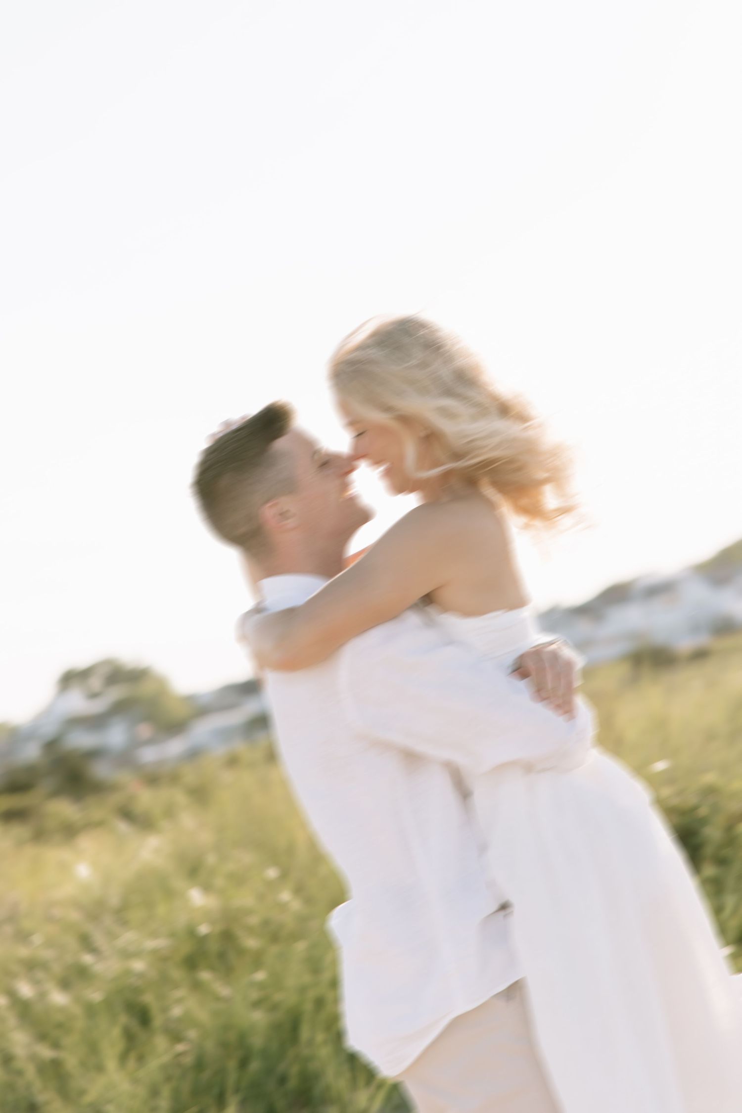 A groom lifts his bride in a sun-drenched field, sharing a romantic moment in elegant white wedding attire.