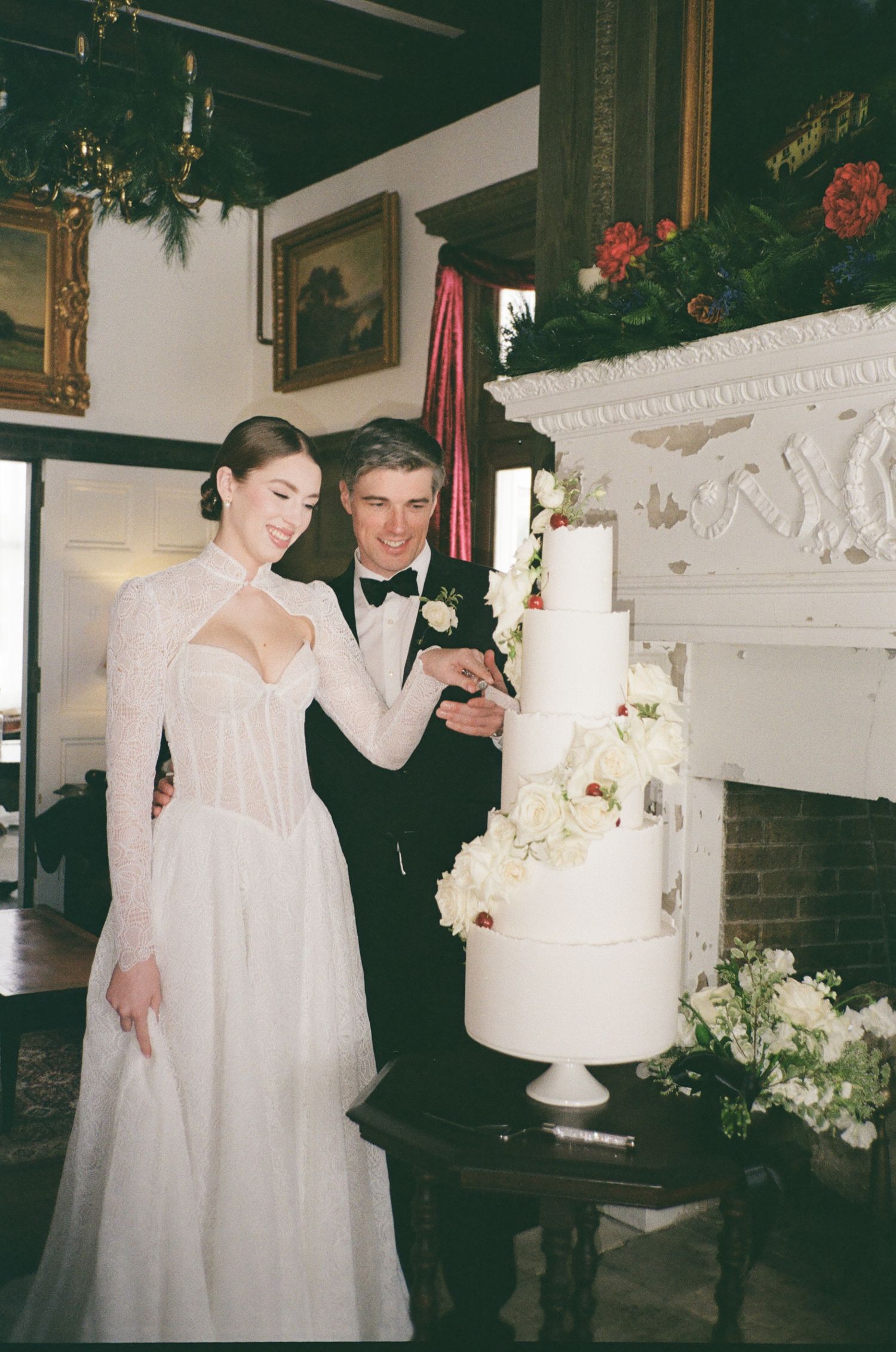 Bride and groom cutting a tall white floral wedding cake beside an elegant fireplace in a classic venue.