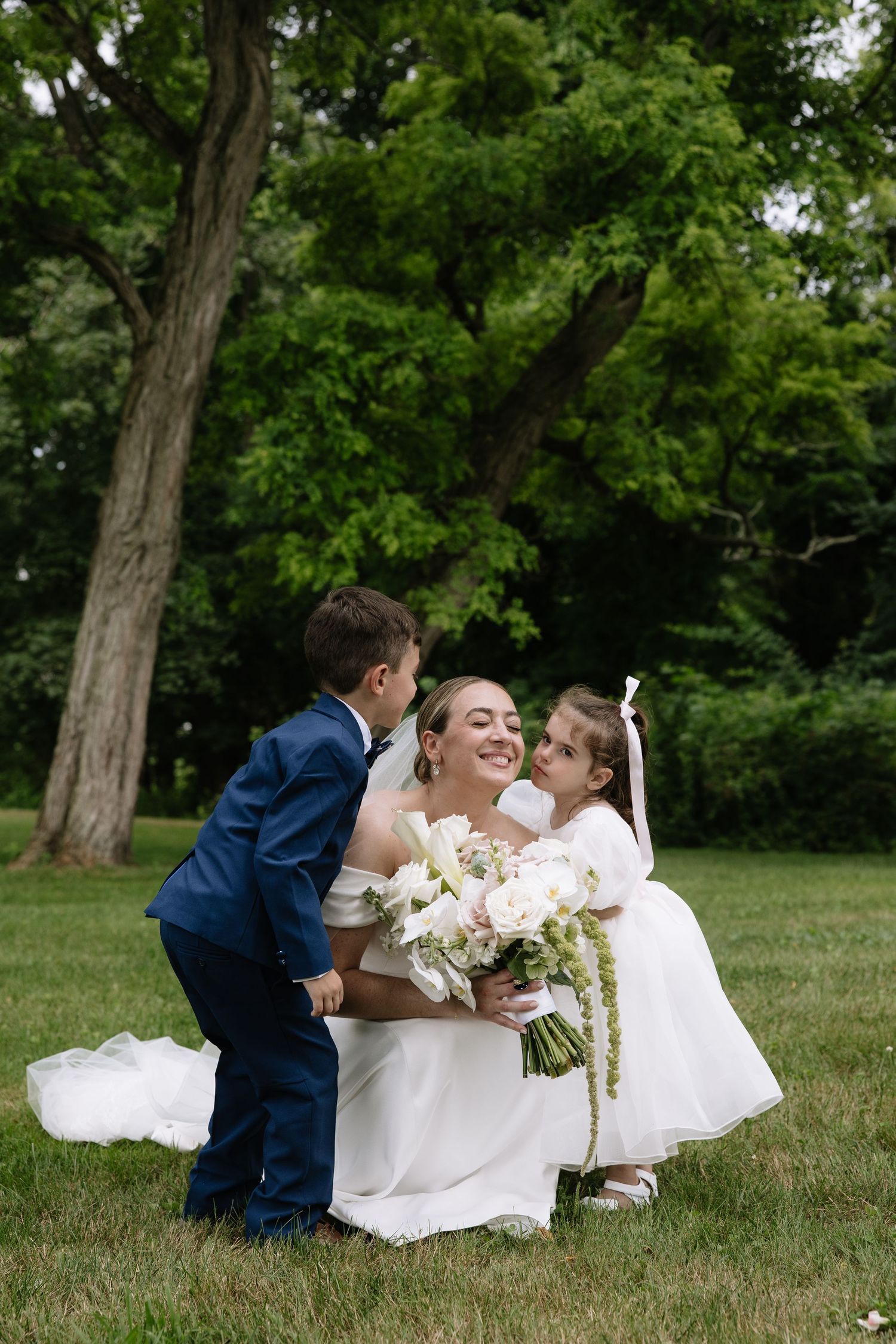 Smiling bride in white gown hugging young ring bearer and flower girl outdoors on green lawn.