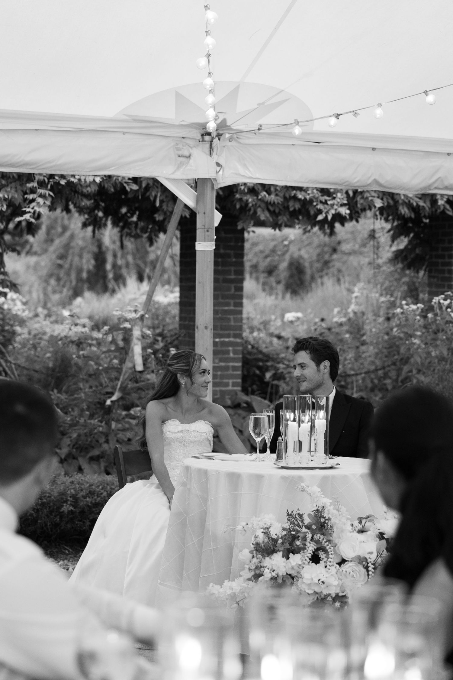 Black and white photo of a bride and groom sitting at their sweetheart table during an outdoor wedding reception.