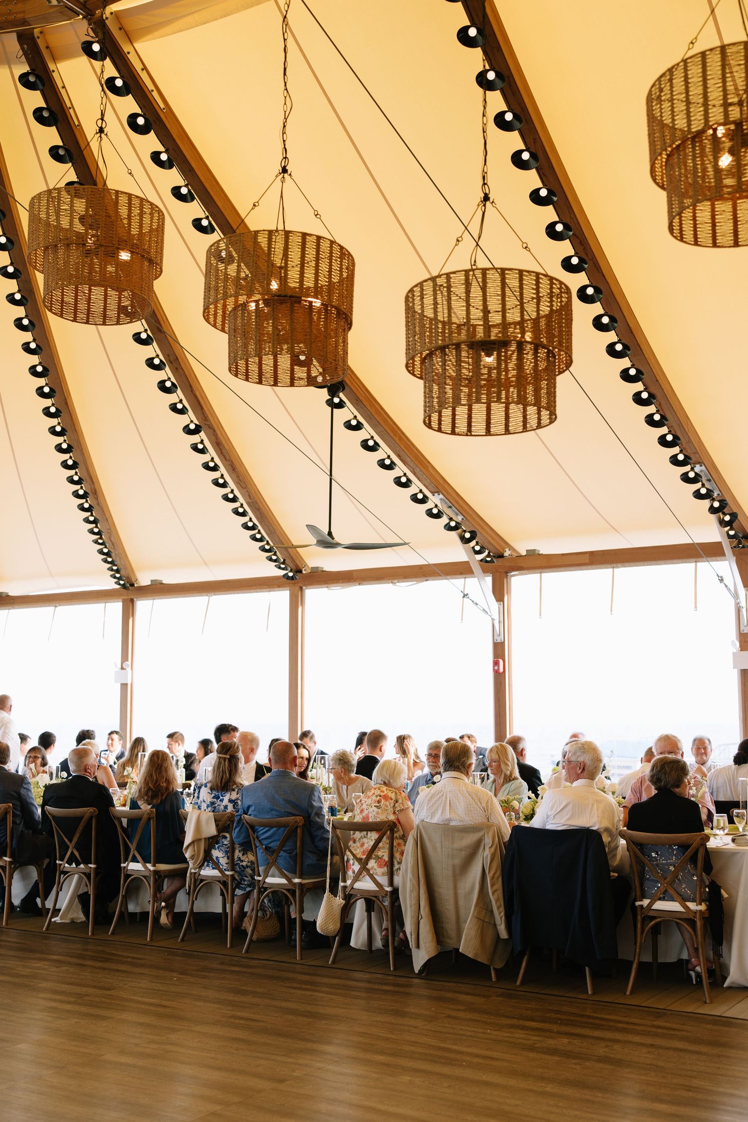 Wedding reception guests dining under a elegant tent with wicker chandeliers and string lights.