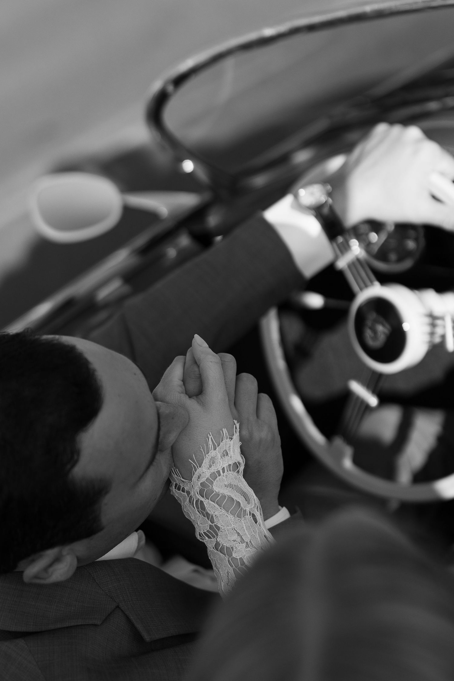 Black and white photo of a couple holding hands in a vintage convertible car, bride wearing lace gloves.
