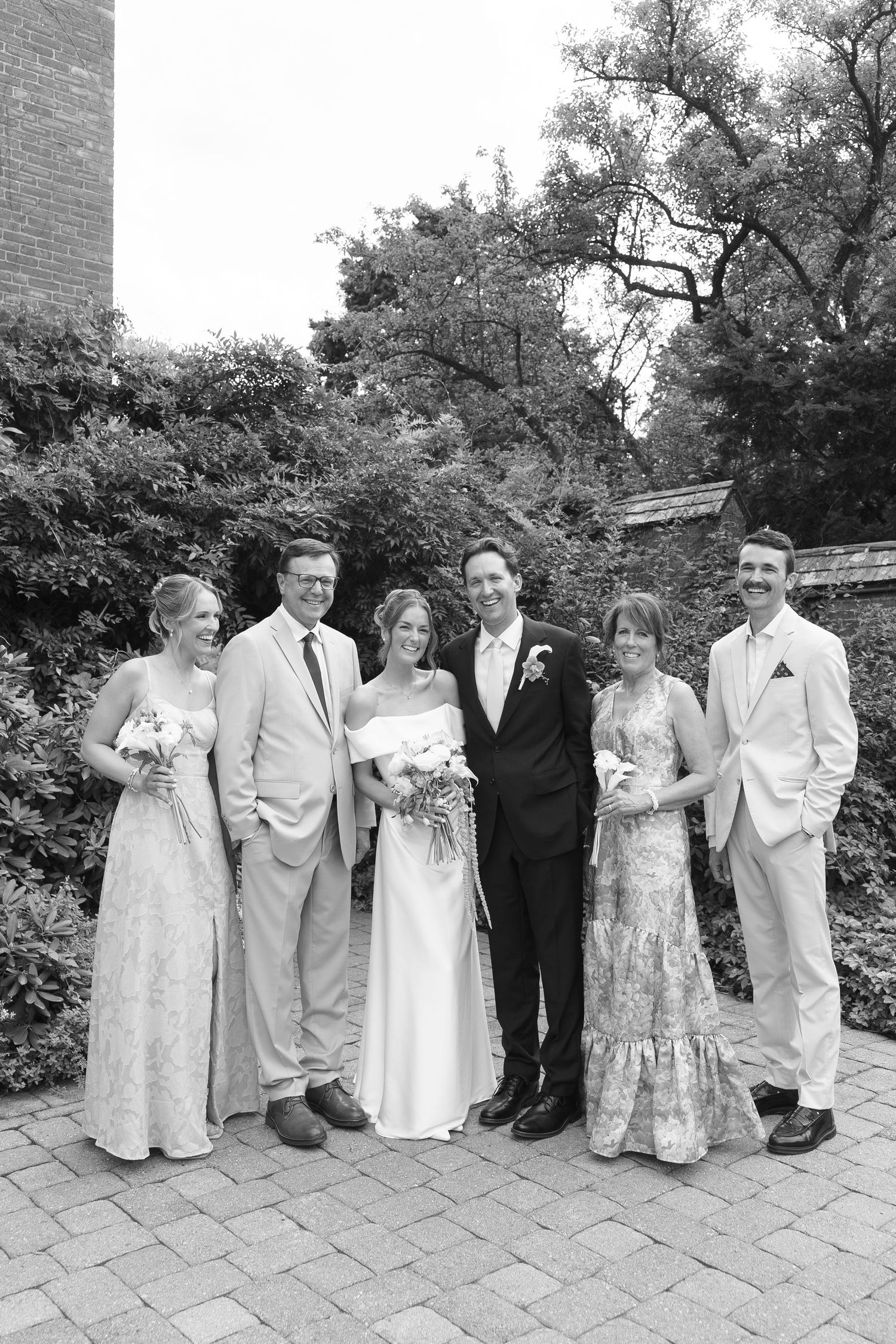 Black and white wedding portrait of a bride and groom surrounded by family in a garden, all dressed up and smiling.