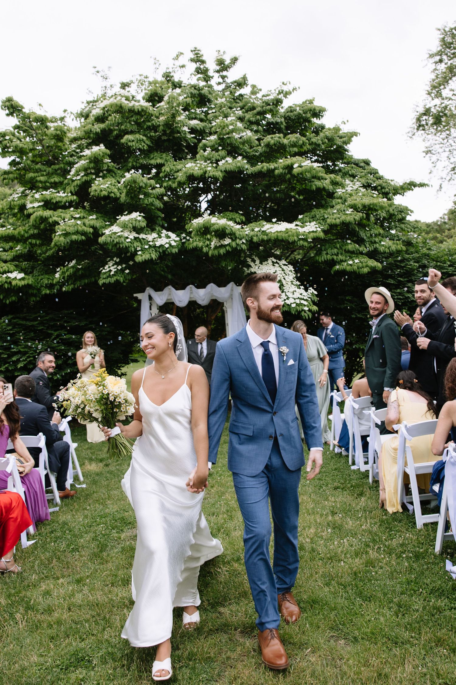 Bride and groom walk hand-in-hand down outdoor aisle after wedding ceremony, surrounded by cheering guests.