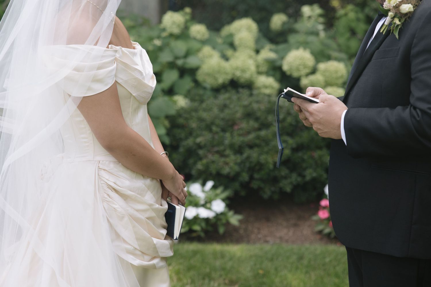 Bride and groom exchanging vows outdoors, holding notes during wedding ceremony with lush green hydrangeas in background.
