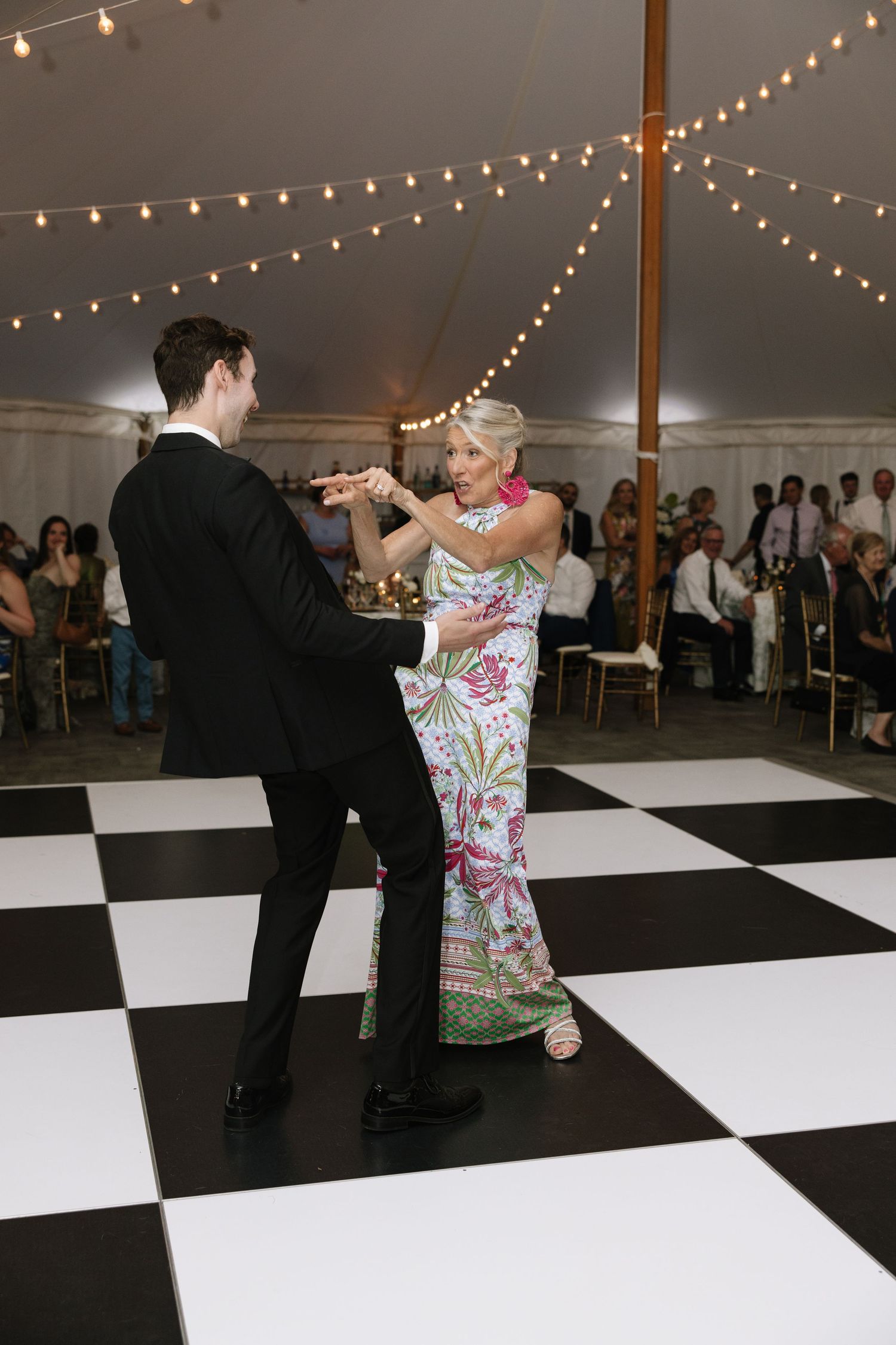 Groom dances with elegantly dressed woman in floral gown on black and white checkered dance floor under a reception tent.