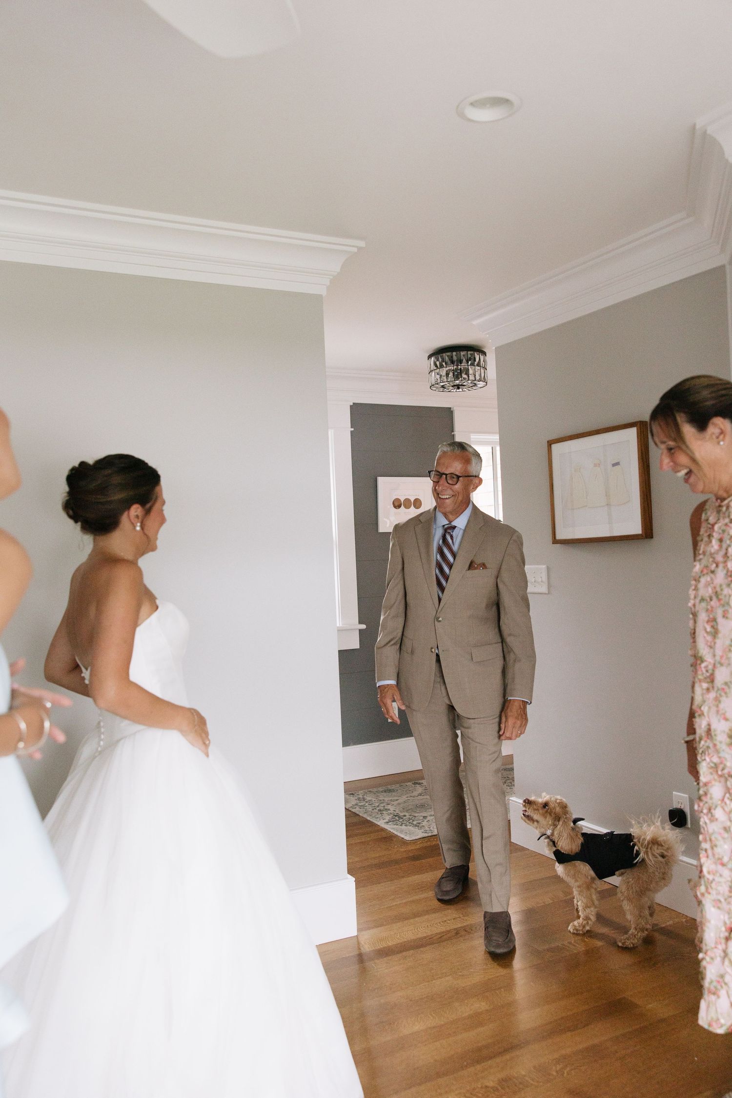 Father sees bride in wedding dress for first time, sharing emotional first look moment with small dog in tuxedo nearby.