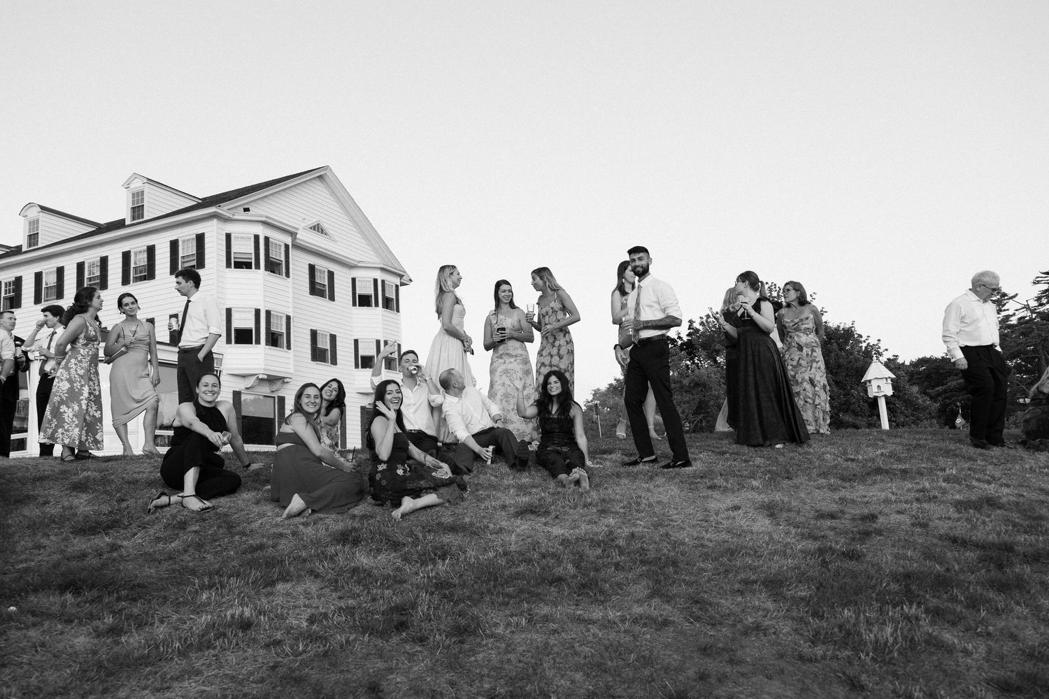 Wedding guests gathered on a lawn at dusk in front of a large white colonial estate in black and white photo.