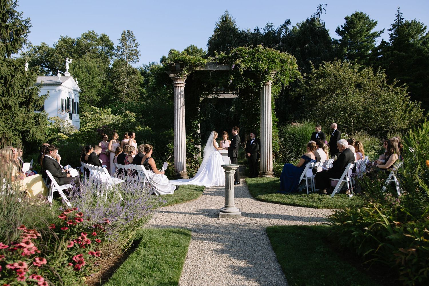Garden wedding venue with stone pillars and white chairs along a winding path.