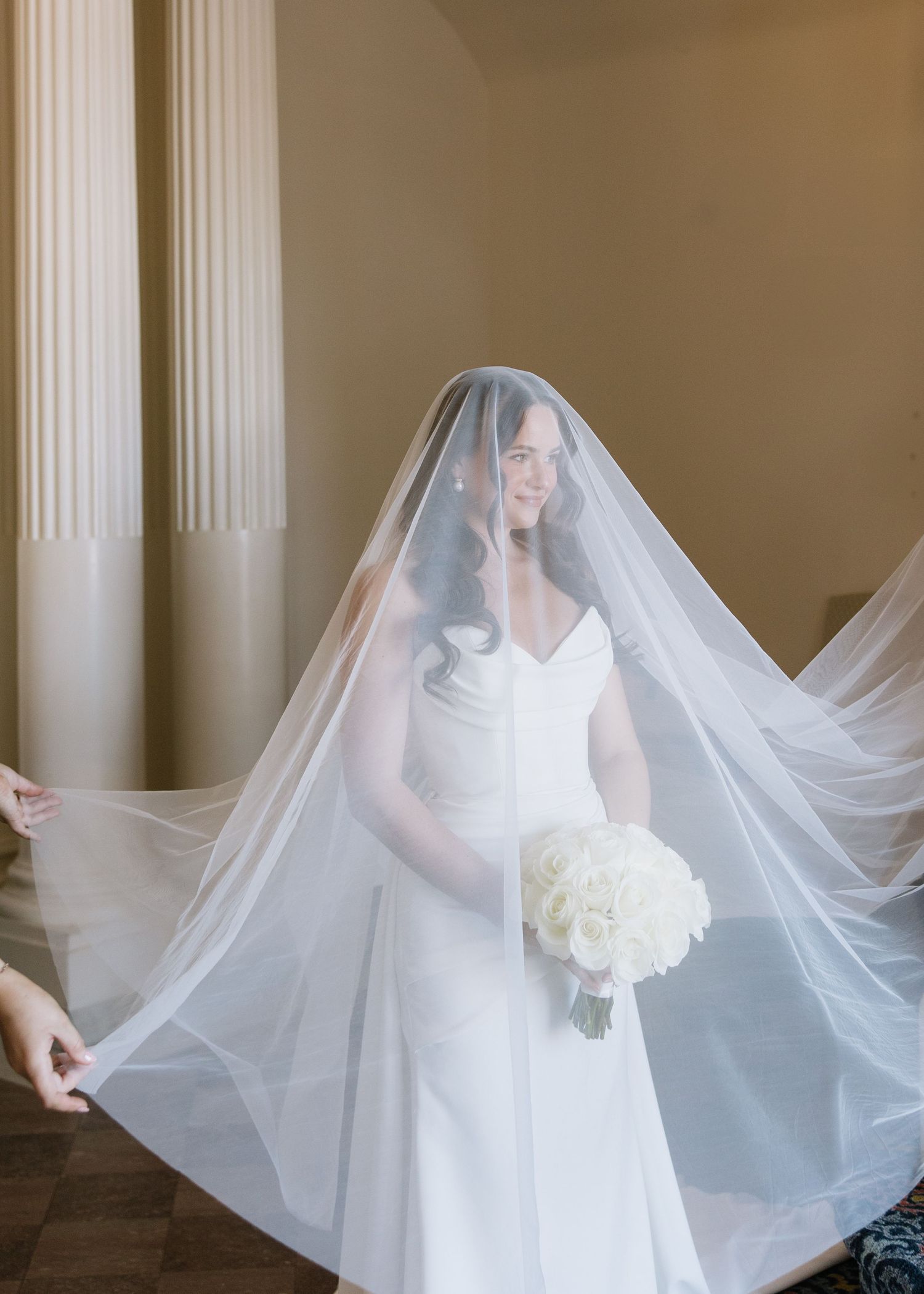 Bride in white dress holding bouquet with flowing veil in elegant interior.