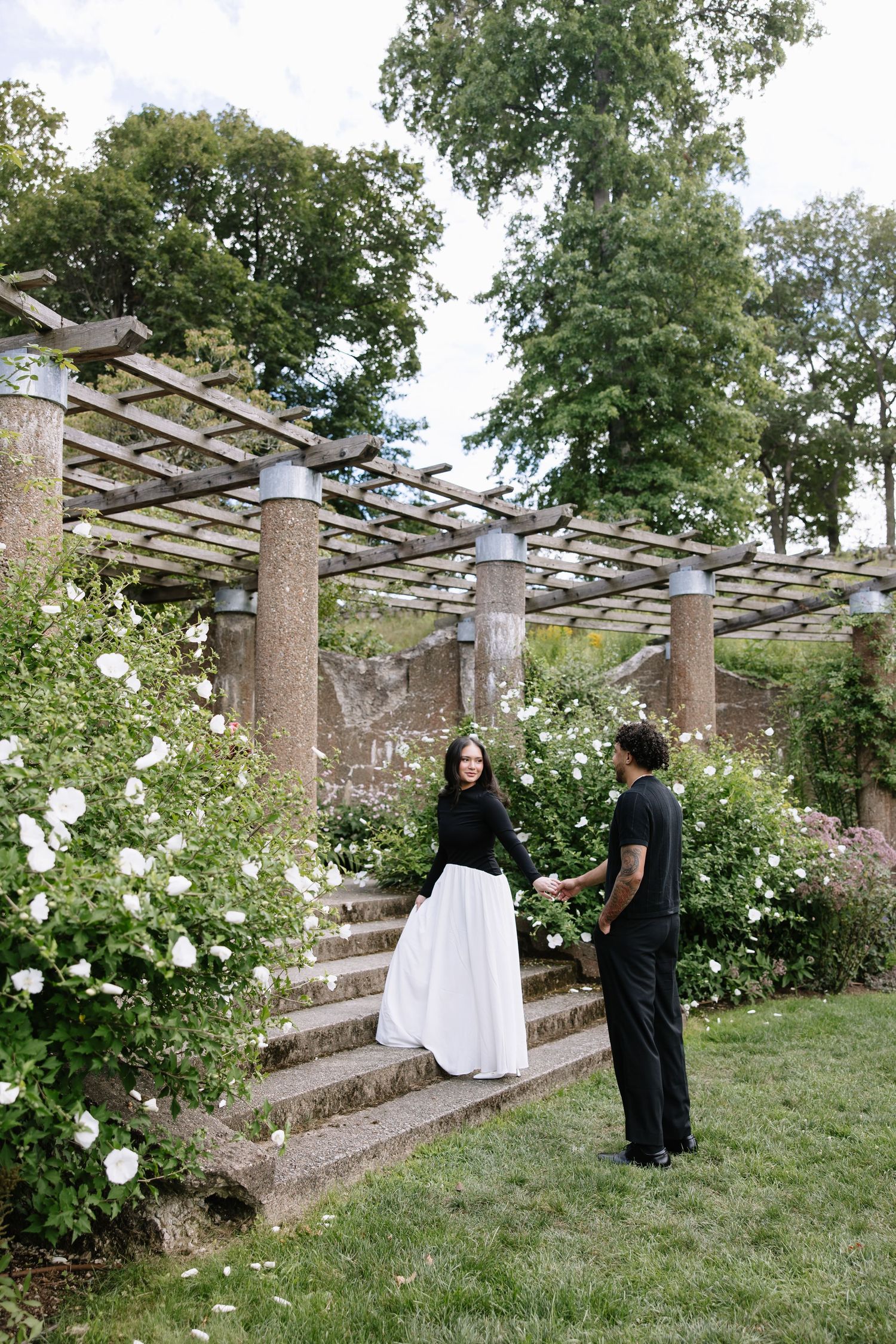A couple walks through a garden pergola surrounded by white climbing roses and stone columns.