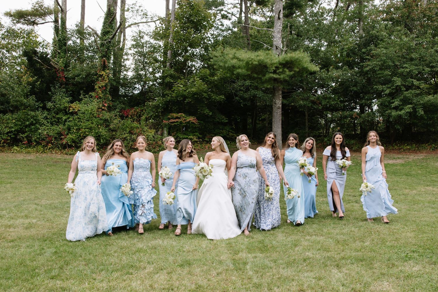 Bridesmaids in light blue dresses pose together on a grassy lawn for wedding photos.
