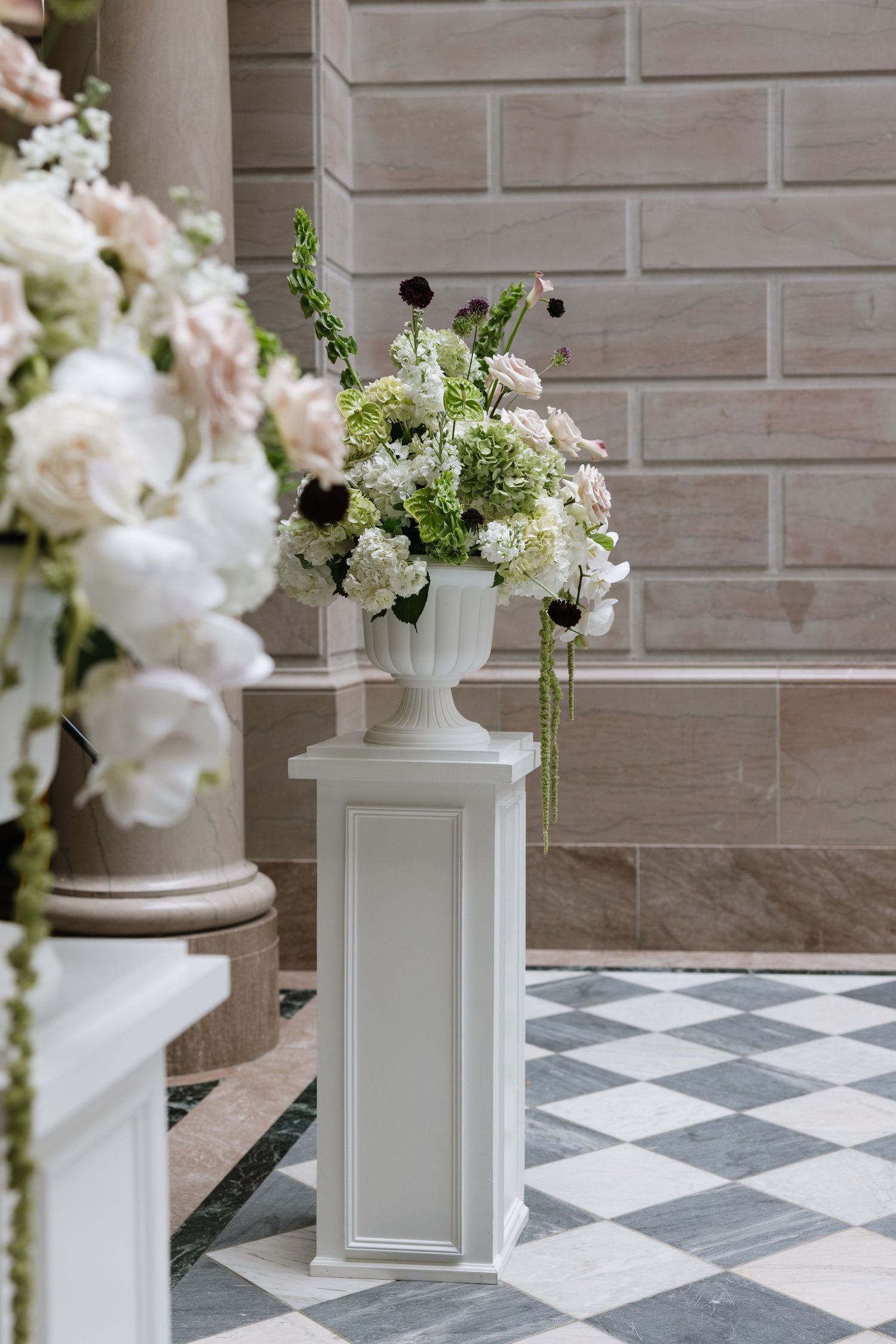 White urn vase with lush green and white floral arrangement on pedestal in elegant wedding venue.
