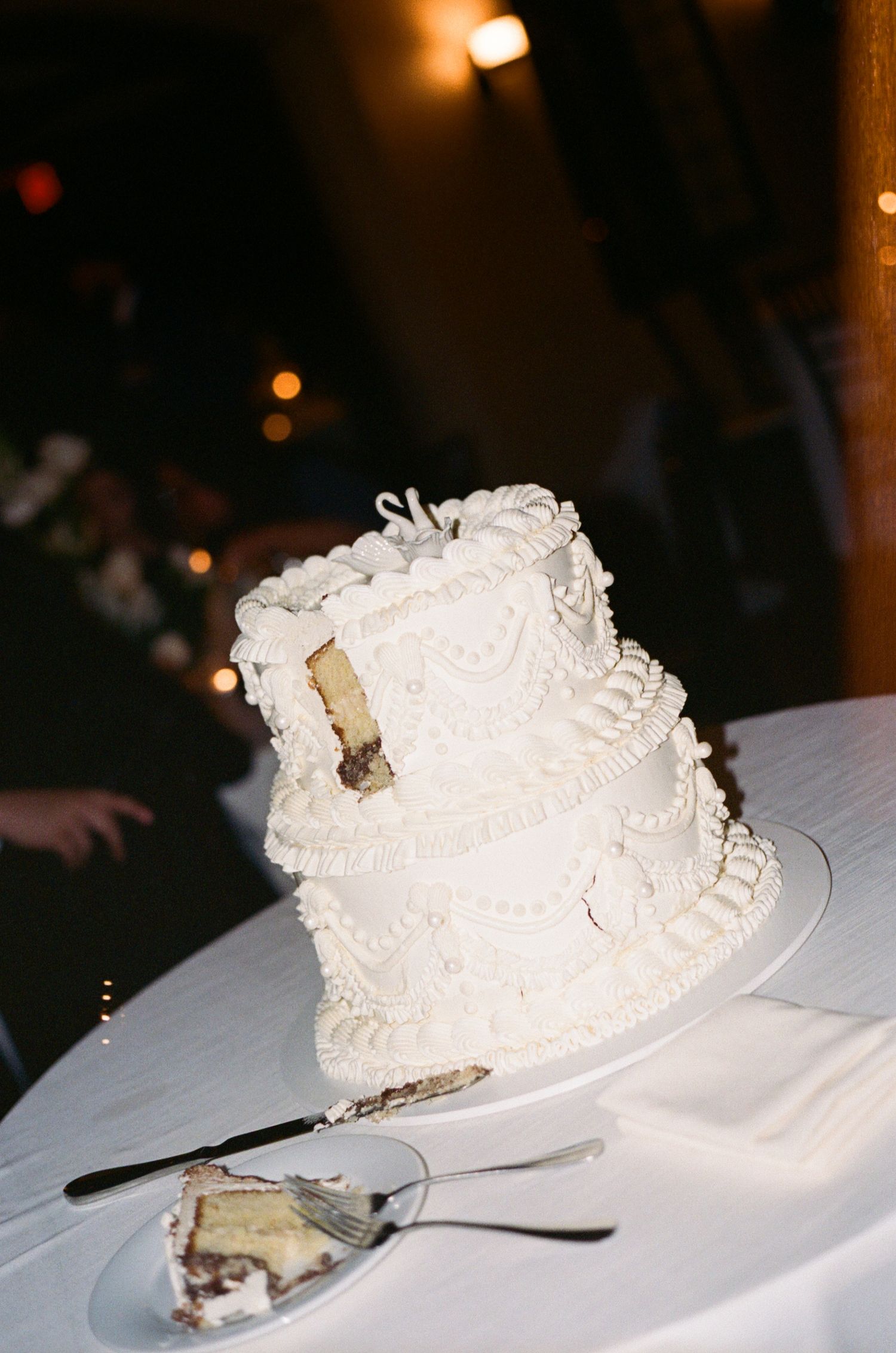Elegant three-tier white wedding cake with intricate piped frosting details, partially cut, displayed on a white tablecloth.